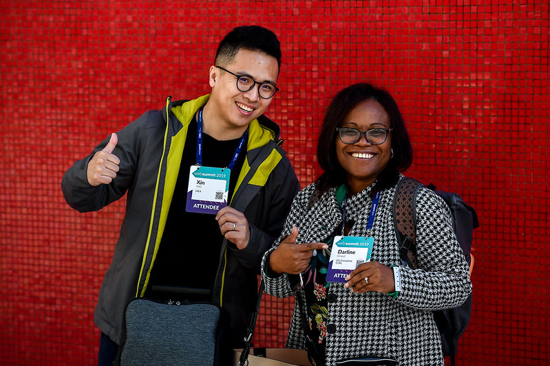 Two smiling people stand in front of a tiled wall. One has a small bag at their waist. They are giving a thumbs up with one hand and holding an event accreditation lanyard – which they're wearing around their neck – with the other. The other person is wearing a backpack. They're holding their event accreditation lanyard – which they're wearing around their neck – with one hand, and pointing at it with the other.
