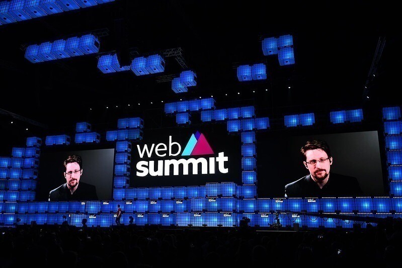A close-up of Web Summit Centre Stage. There are large screens on either side of the Web Summit logo, each showing Edward Snowden.