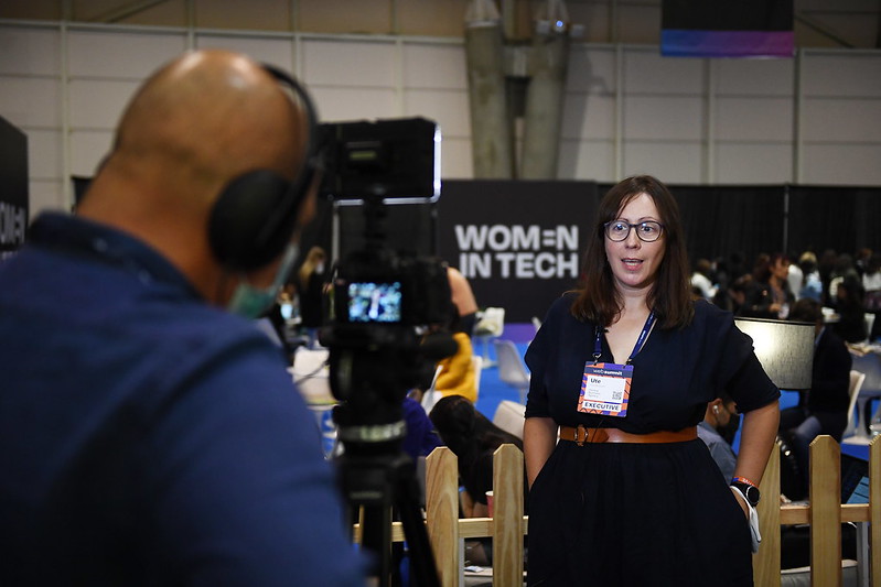A person stands with their hands in their pockets in front of a picket fence. The fence runs in front of an area where several people are seated. A sign in this area reads 'Women in tech'. The person is speaking to a video camera, and appears to be giving an interview.