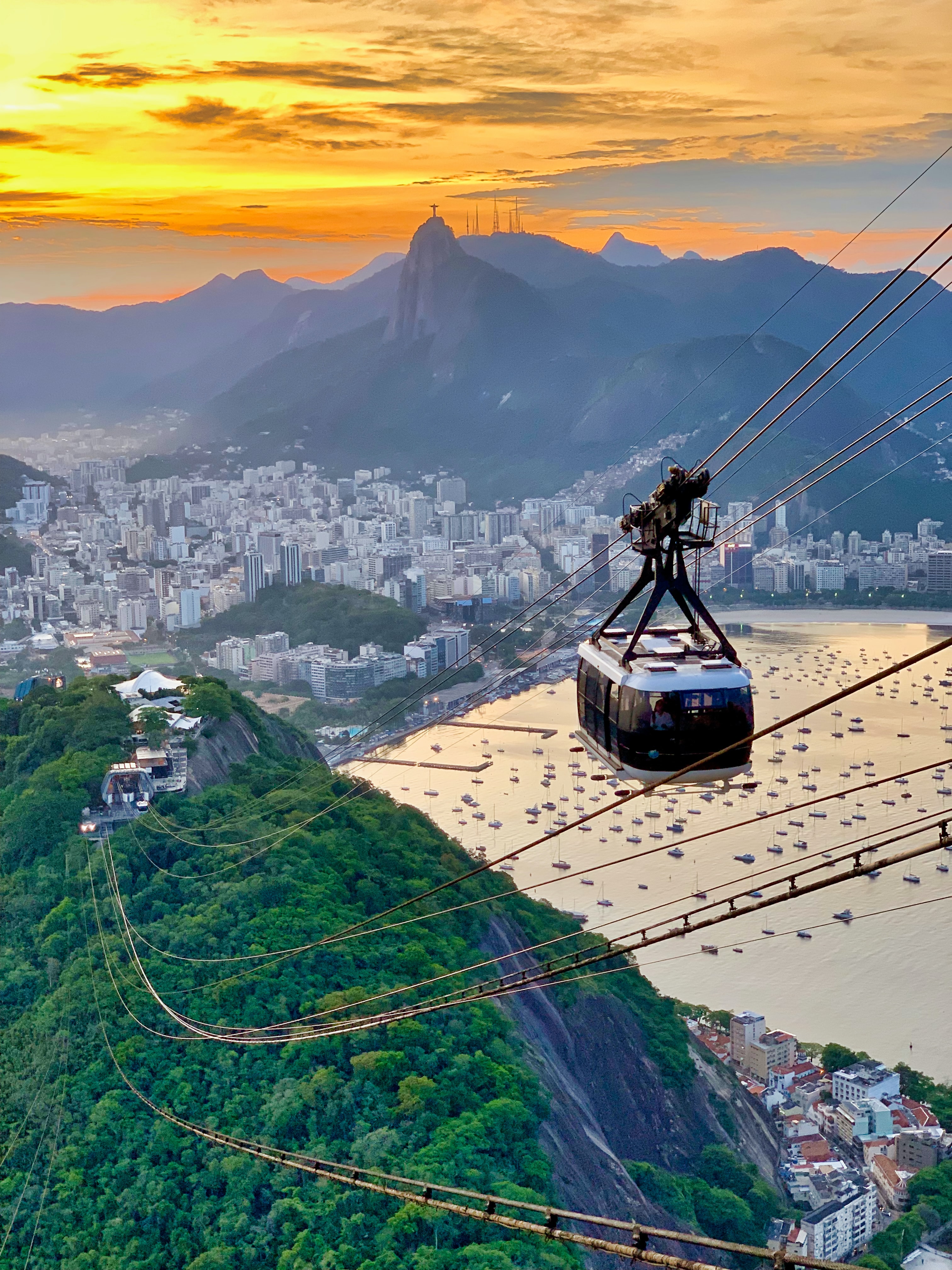A glass-walled cable car descends Rio's Sugarloaf Mountain at sunset, travelling towards a rocky, forested hill. The city of Rio is visible in the background, surrounded by mountains.