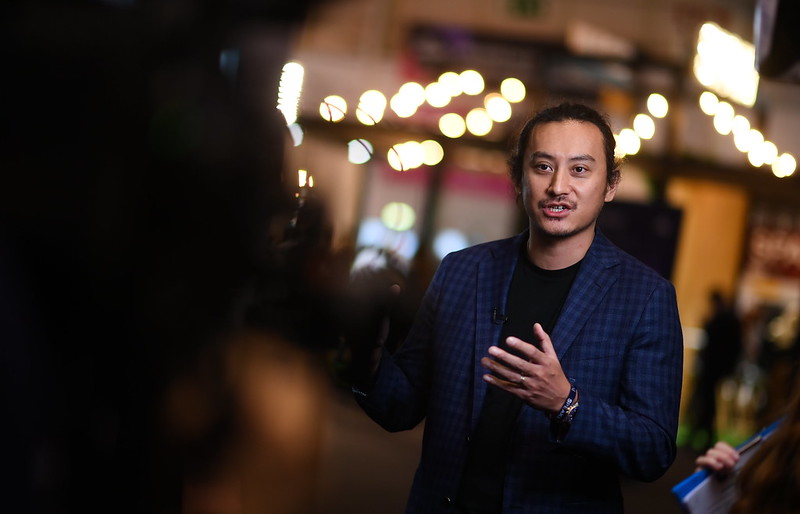 A person (Junta Nakai of Databricks) wearing a suit jacket over a plain crew-neck tshirt. Junta is being interviewed. A video camera is in the foreground, out of focus. Junta is gesturing emphatically and appears to be speaking.