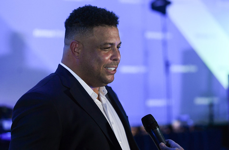 A photograph of Brazilian footballing legend Ronaldo standing in front of a microphone. He appears to be speaking.