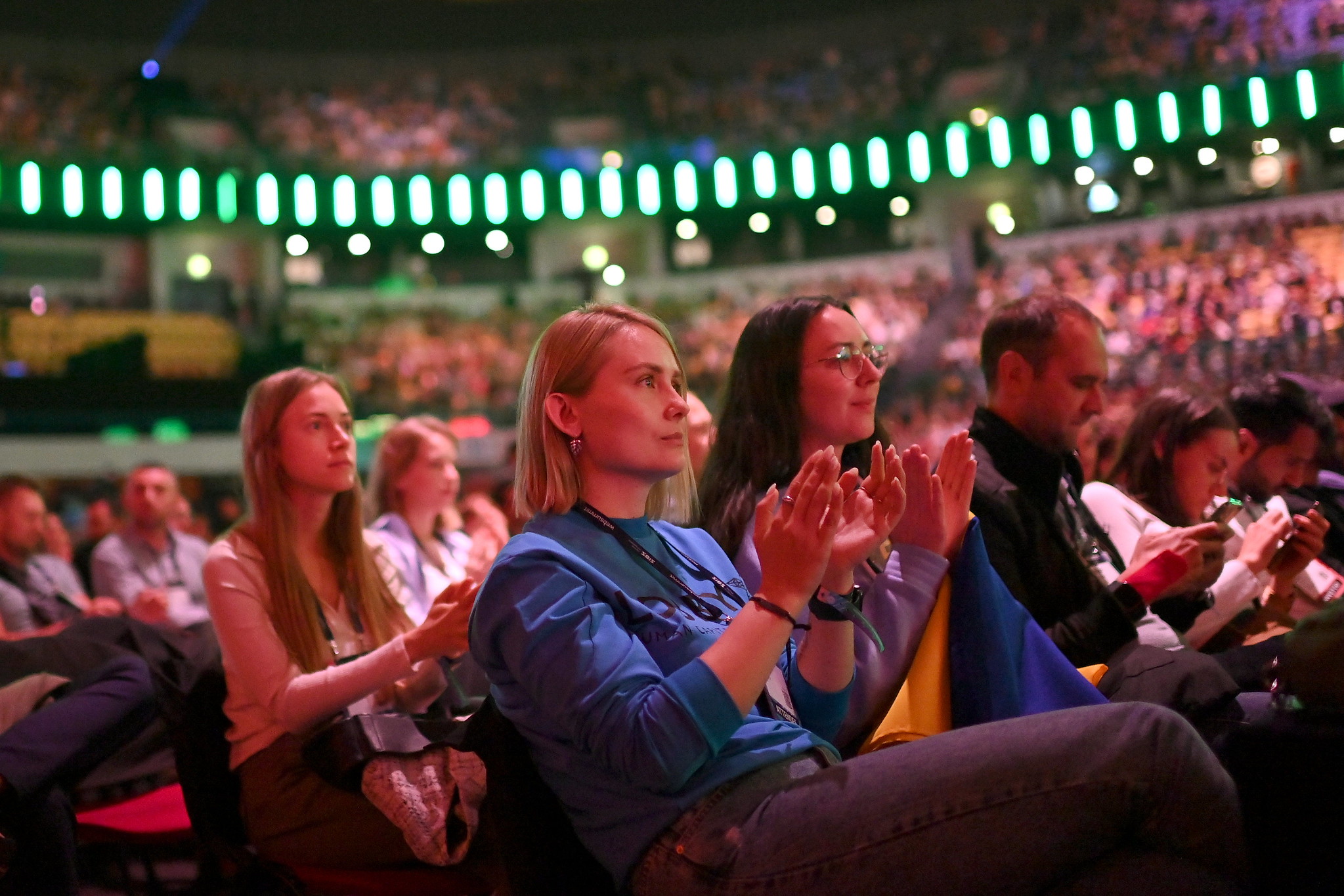 Two women, seated among a vast audience in an arena, applaud while watching a speaker on Centre Stage.