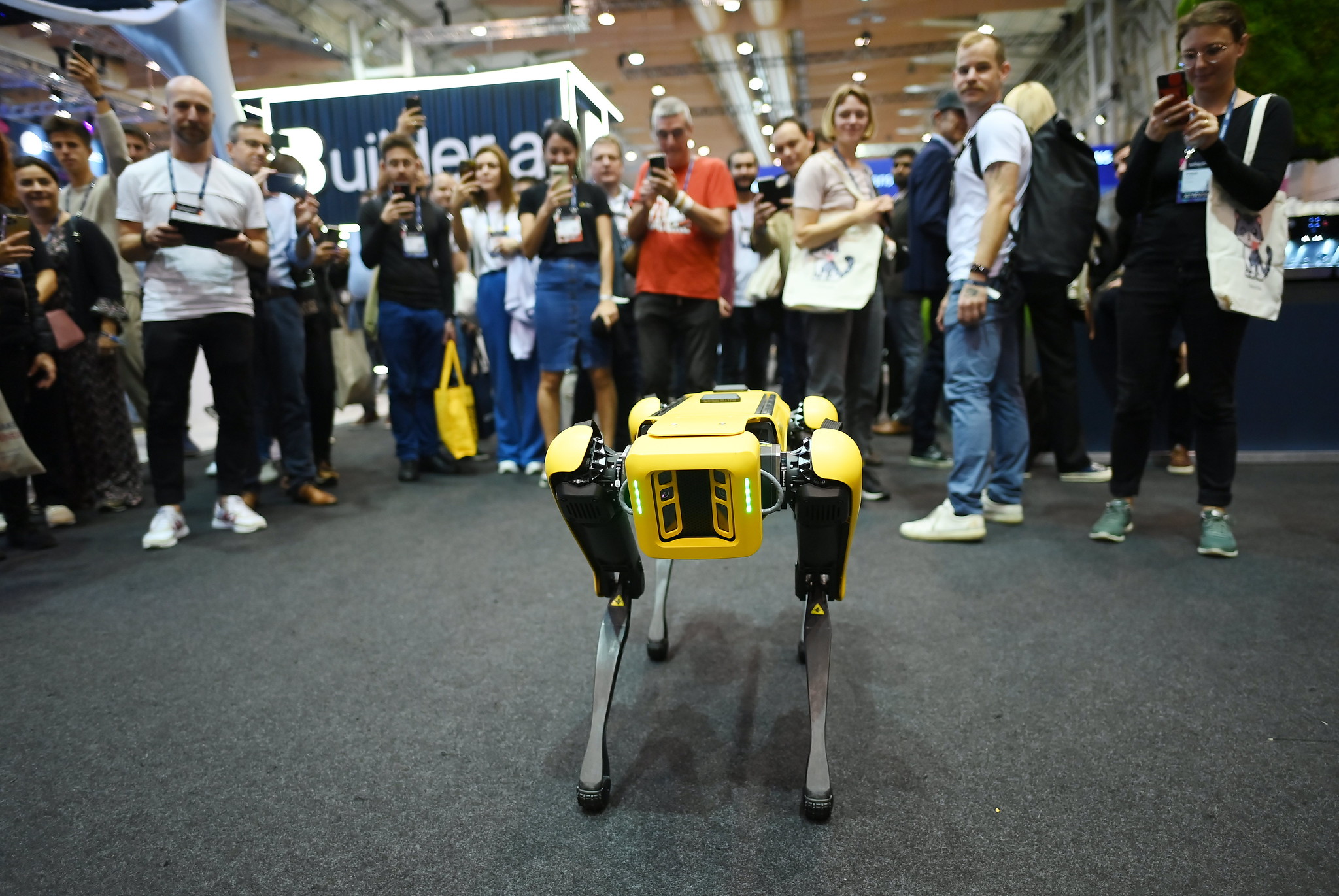 Spot, the Boston Dynamics robot, stands in the foreground. The robot is surrounded by a crowd of people, some of whom are taking photos.