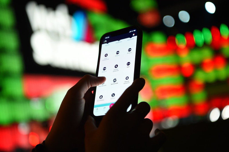 A mobile phone is held in two hands. On the screen of the phone is the Web Summit app. The person holding the phone is perusing the Web Summit schedule. Out of focus in the background is the Web Summit logo on an event stage.