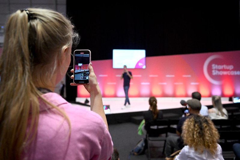 A person in the foreground holds their mobile phone. They're taking a photo of someone on a stage. The stage includes a wall featuring the text 'Startup Showcase'.