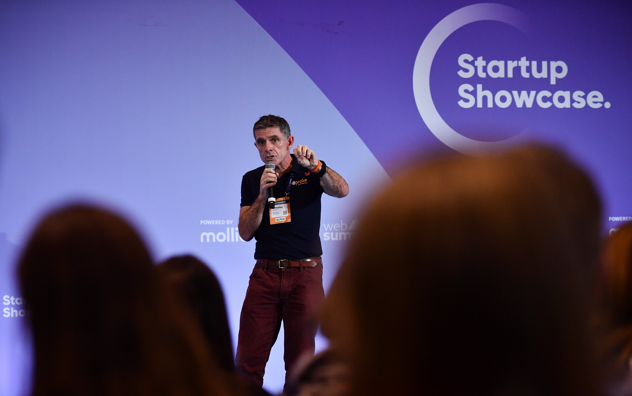 A person stands on a stage holding a microphone and gesturing emphatically. They appear to be speaking. They stand in front of a wall that features the text 'Startup Showcase'. The backs of audience members' heads crowd the foreground.