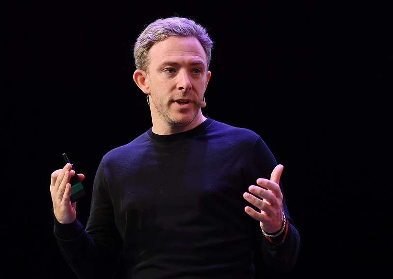 A person (Dfinity founder Dominic Williams) pictured from the waist up. They are gesturing with both hands. A presentation clicker is held in the right hand. They are wearing a headset mic and appear to be speaking.