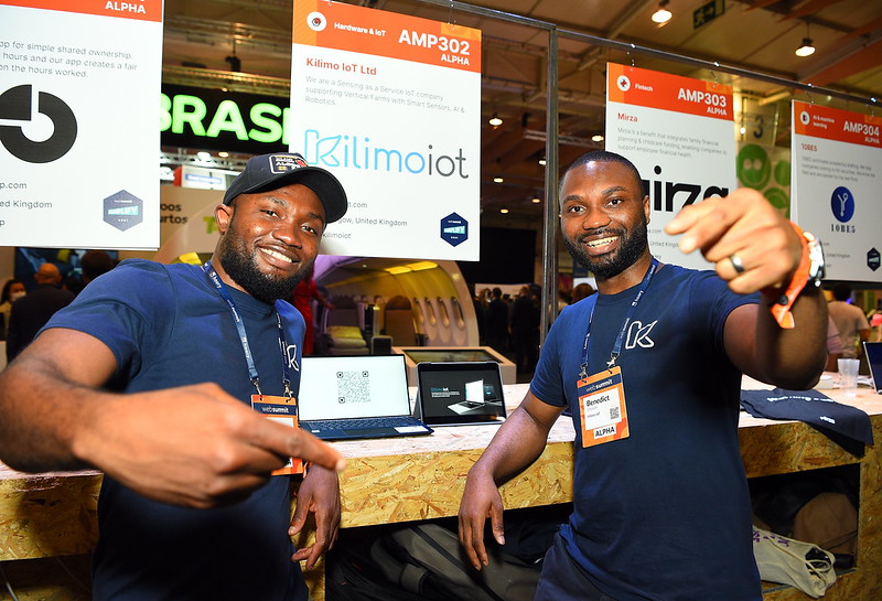 Two people stand in front of a chipboard counter. Above them, placards hang that detail the names, logos and elevator pitches of startups. The two people are smiling and pointing towards the camera. They appear to be representatives of a startup called Kilimo I.O.T