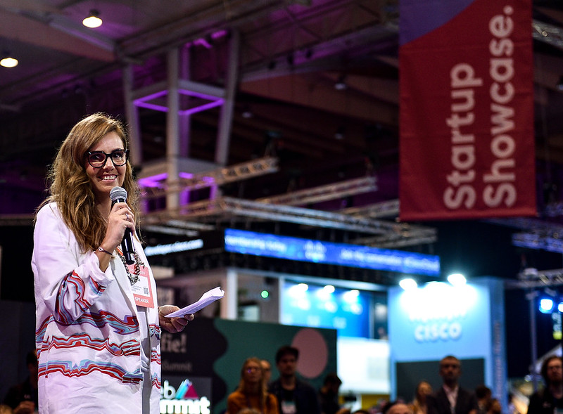 A smiling person stands to the left of the image. They're holding a microphone and a piece of paper, and appear to be talking. In the background are several exhibition stands, as well as a hanging banner reading 'Startup Showcase'.