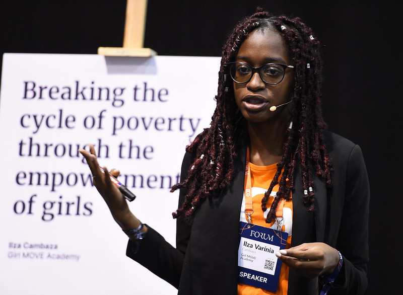 A person stands in front of an easel that holds a sign reading 'Breaking the cycle of poverty through the empowerment of girls'. The person is wearing a headset mic and gesturing with their right hand. They appear to be speaking.