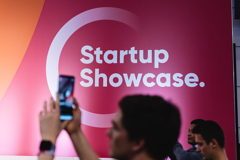 A person holds up a mobile phone in front of a wall featuring the Startup Showcase logo