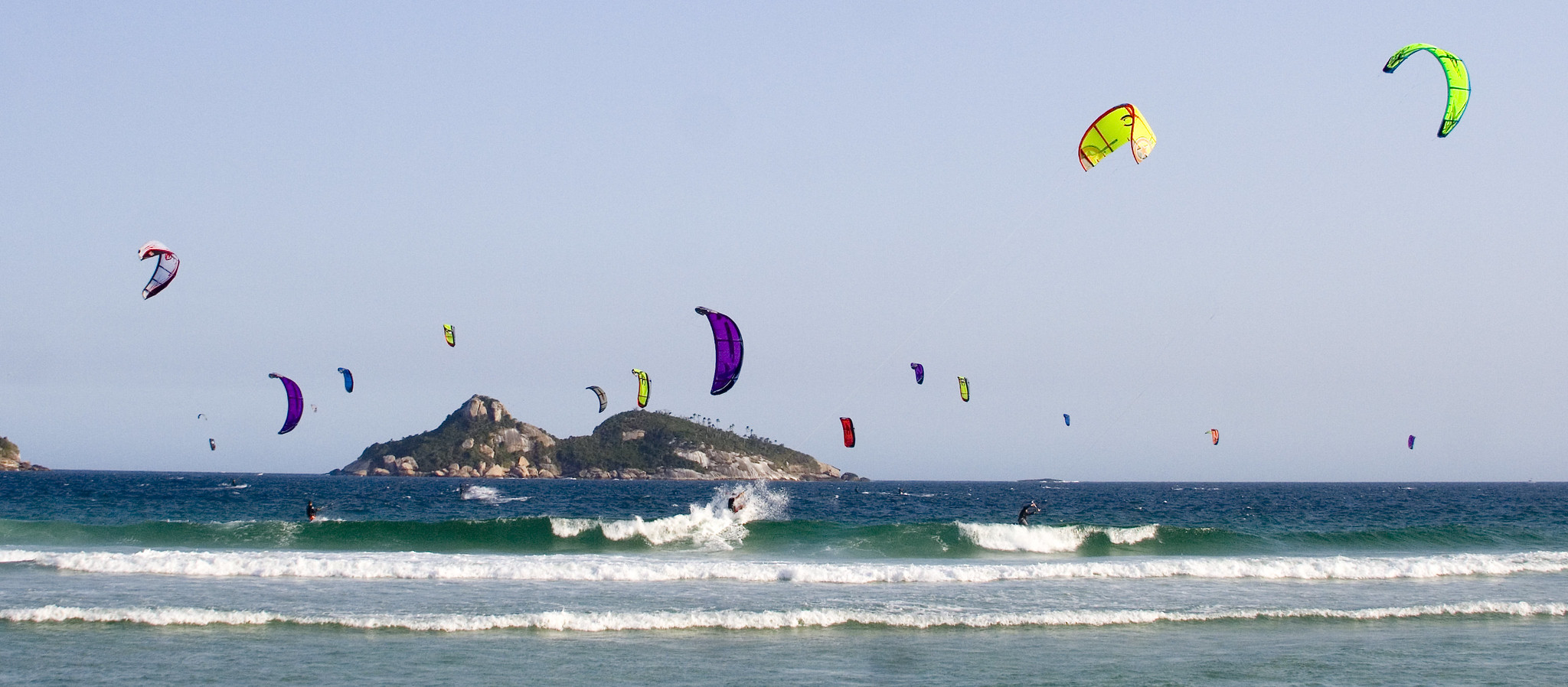 Low gentle waves appear in the foreground, spread across the width of the photo. Larger waves break behind them. Two kitesurfers crest the largest wave. The sky is dotted with kitesurfing kites. In the background, a rocky, forested island is visible on the horizon.