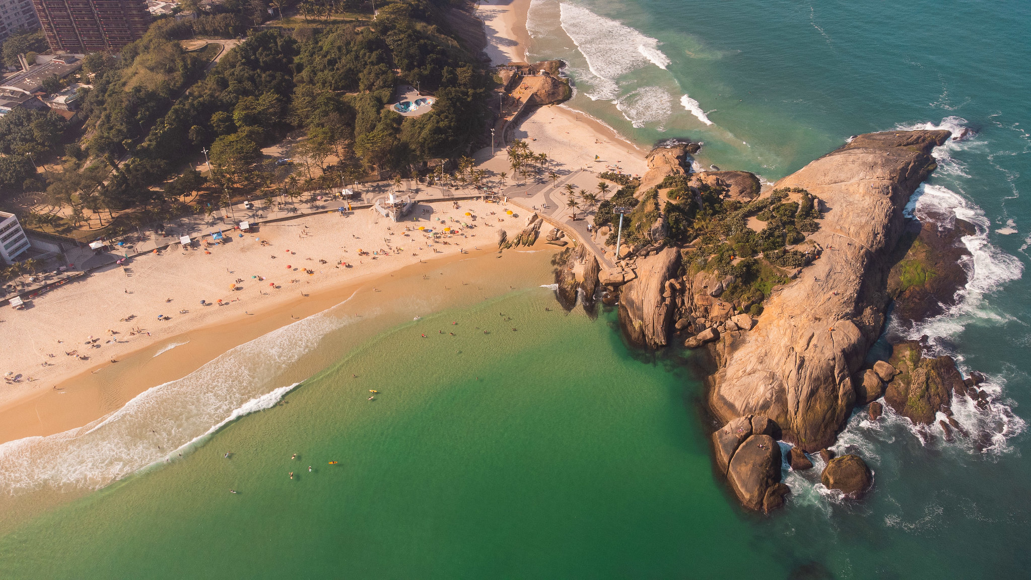 An aerial view of a stretch of sand lining a bay. A large cluster of rock abuts the beach at the point where it meets the next bay. Image: Riotur