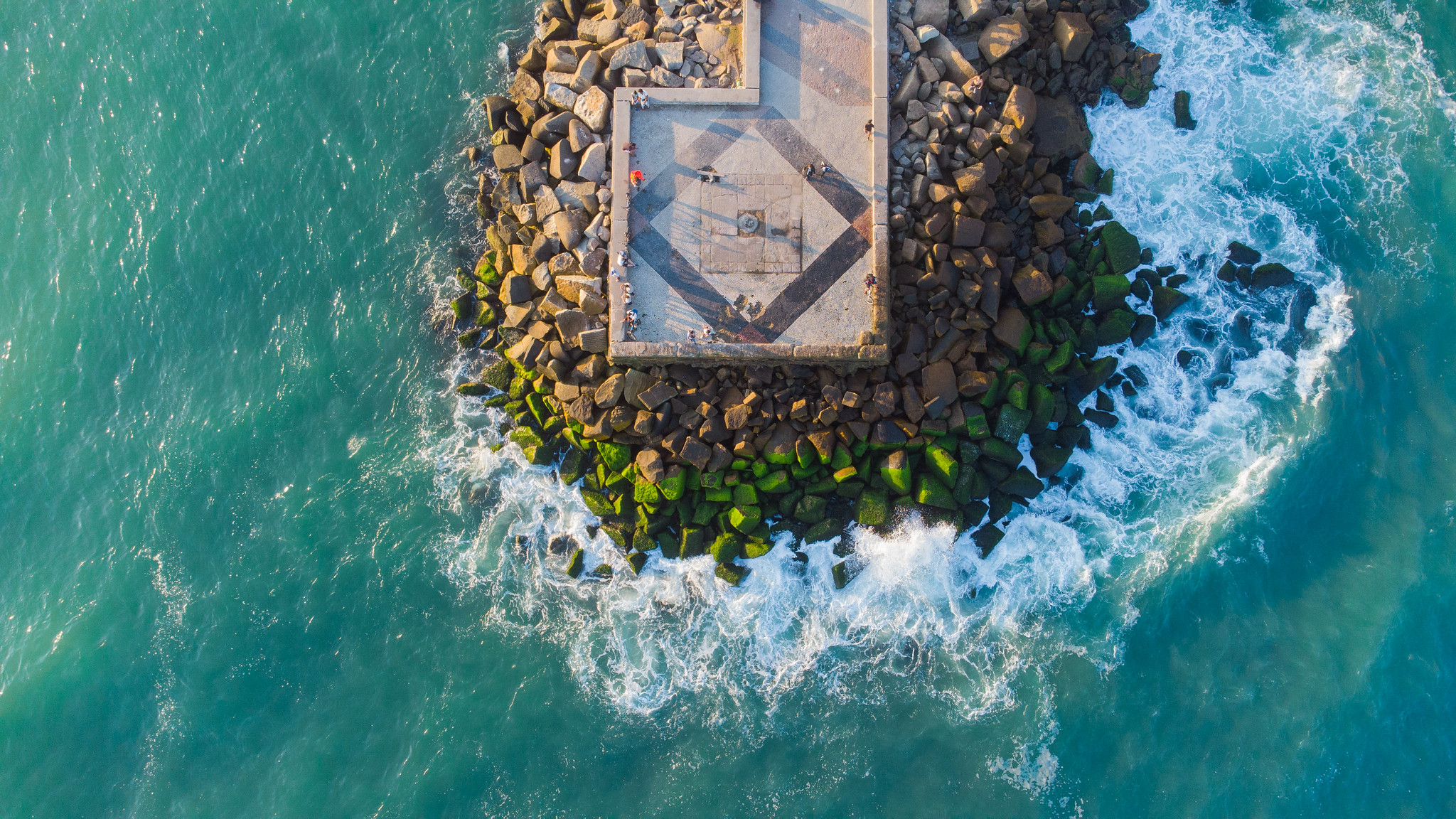A concrete pier ends in a small square. The pier is built on top of a mound of rocks. It's surrounded by water that is frothing against the rocks. Image: Rafael Catarcione/Riotur