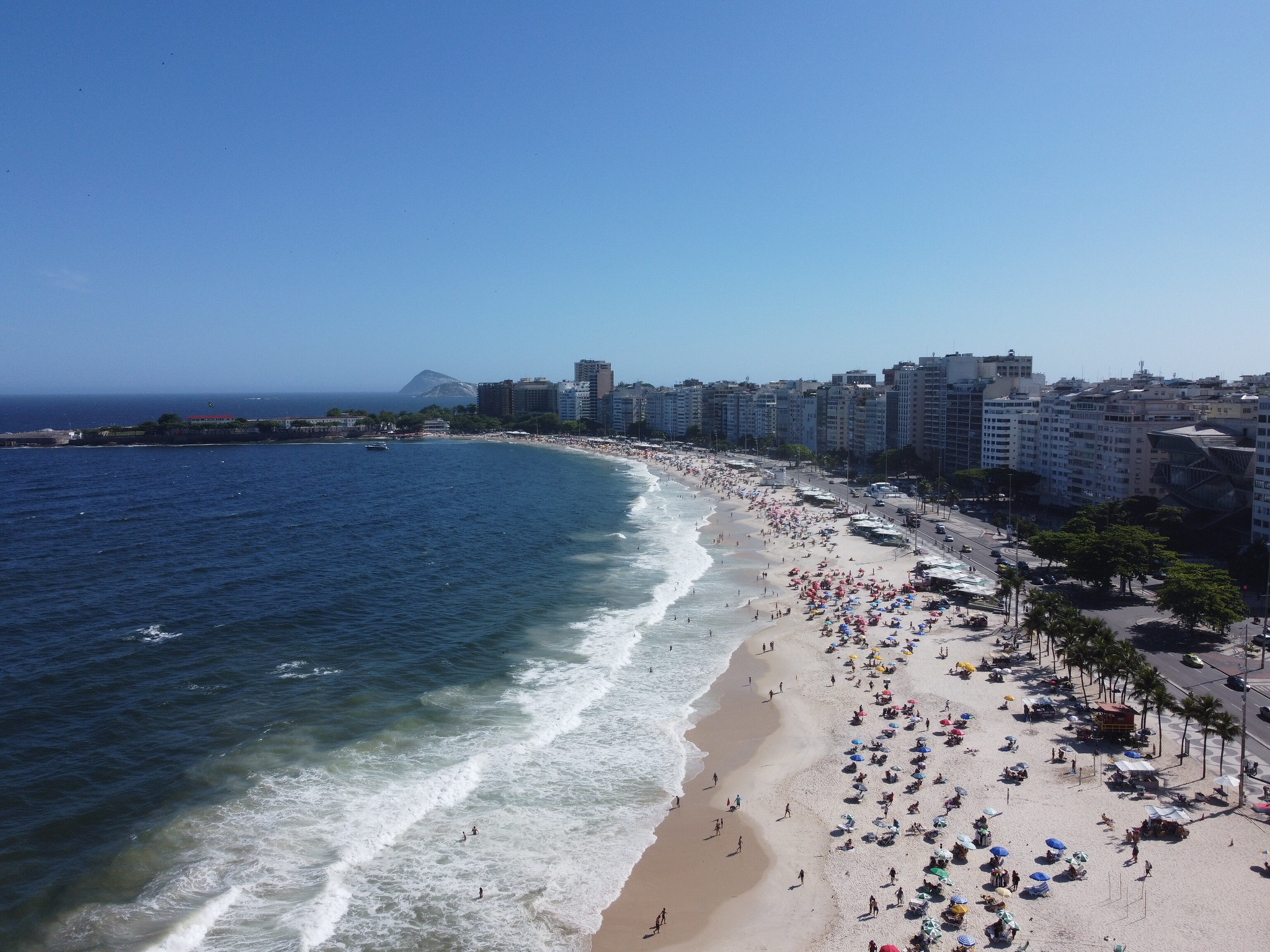 An aerial view of Copacabana beach. A wide strip of sand curves along a bay. The beach is busy with people, but not crowded. To the left of the sand is the sea, with waves lapping the sand. To the right of the sand is a road and, on the other side of the road, a row of multistory buildings. Image: Rafael Catarcione/Riotur.