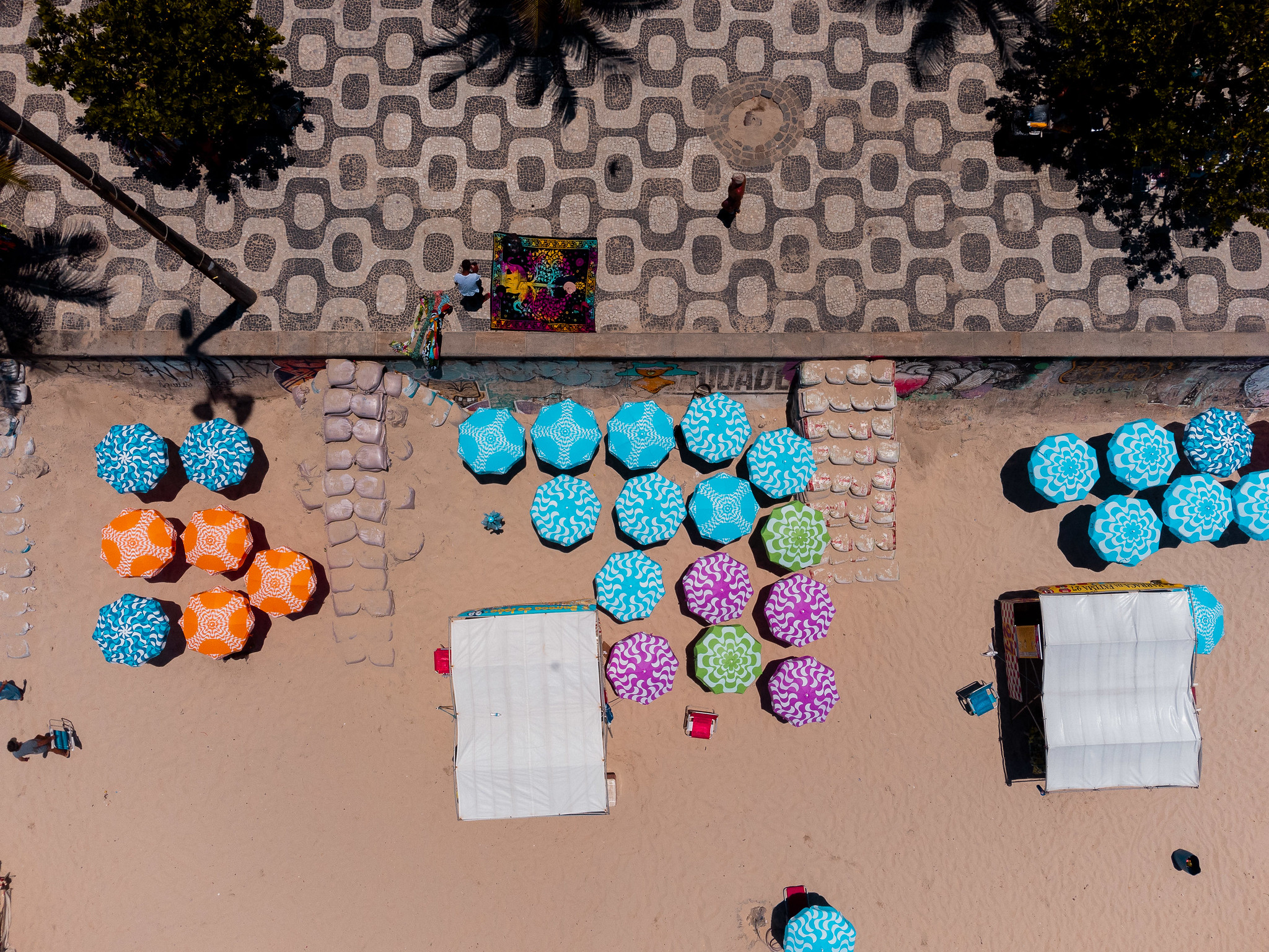 Aerial view of a beach where it meets a tiled walkway. There are clusters of beach umbrellas dotted around the sand.