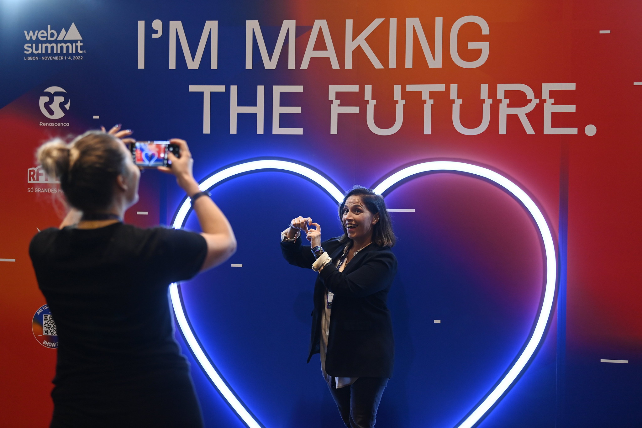A person makes a heart shape with their hands in front of a wall with text that reads 'I'm making the future'. Another person, whose back is to the camera, is taking a photo using a smartphone.