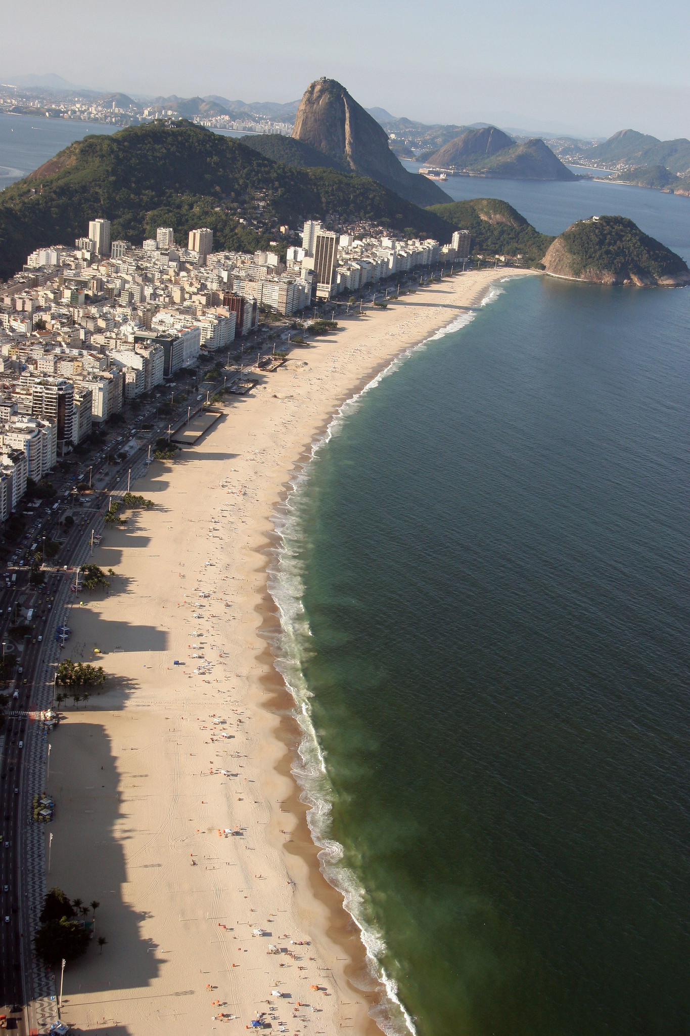 An aerial view of Copacabana beach. A curved stretch of sand reaches towards a large rocky hill. Tightly packed building hug the sand on the left, while the sea laps the beach on the right. Foamy waves appear where the sea meets the sand. Image: Ricardo Zerrener/Riotur