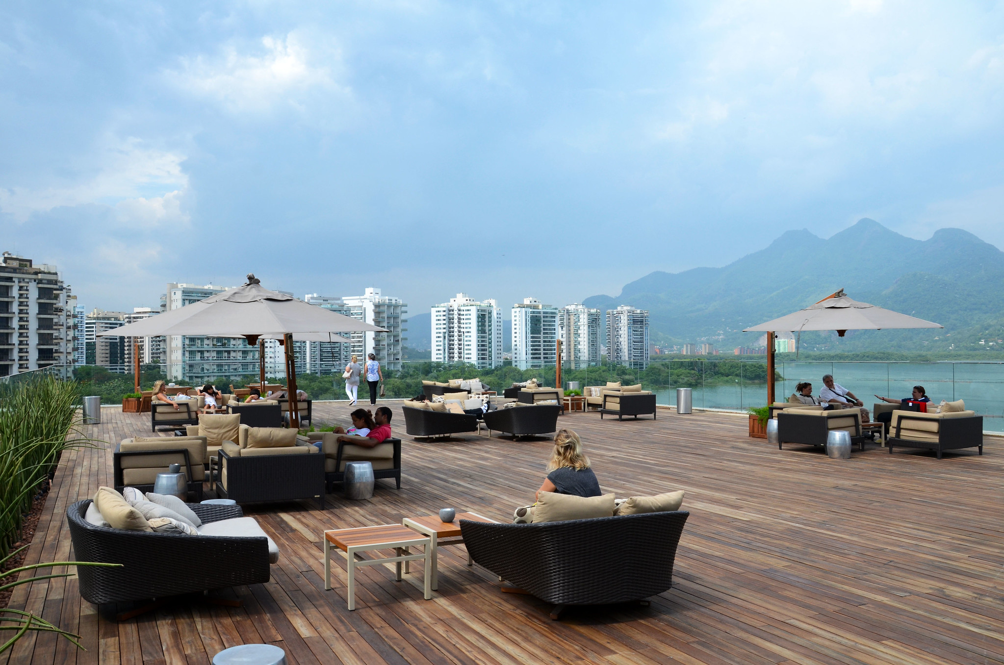 A rooftop relaxation area. Wood-panel flooring stretches the length of the rooftop, which is surrounded by a glass barrier. Dotted around the rooftop are coffee tables, low basket-weave lounge chairs, and sun umbrellas. From the rooftop can be seen multi-story buildings, forested mountains and Lake Tijuca. Image: Alexandre Macieira/Riotur