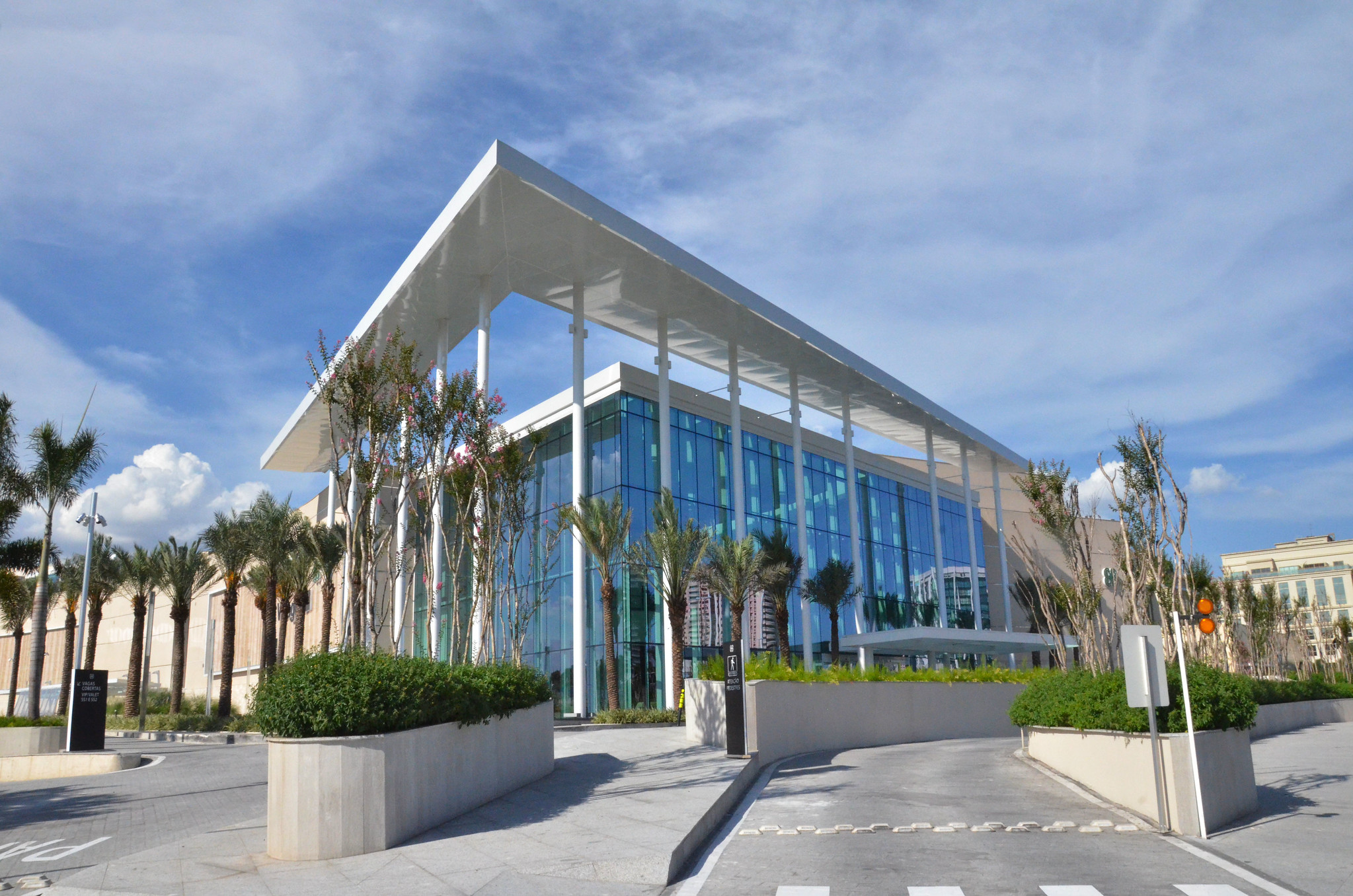 Exterior entranceway of the Village Mall in Barra Tijuca. A glass-fronted building. A wide sun shelter supported by pillars extends around the entranceway separate from the rest of the building. There are two roadways that pass along the front of the building and to the left of the entranceway. Pedestrian walkways are dotted with trees and cement planters.