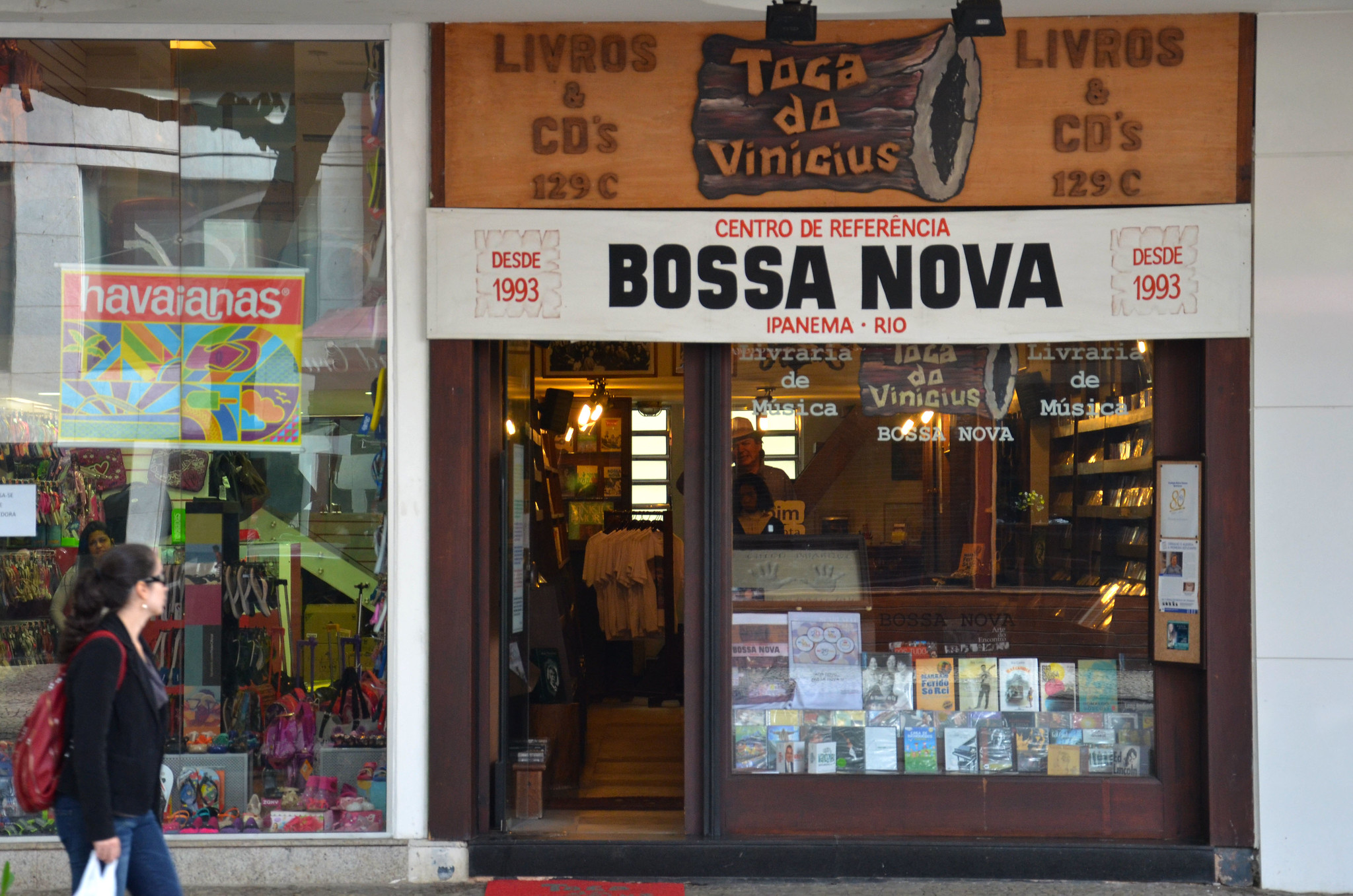 A person walks by a store front. Over the door, a sign reads 'Bossa Nova' and another reads 'Toca do Vinicius'. The store appears to sell books and CDs.