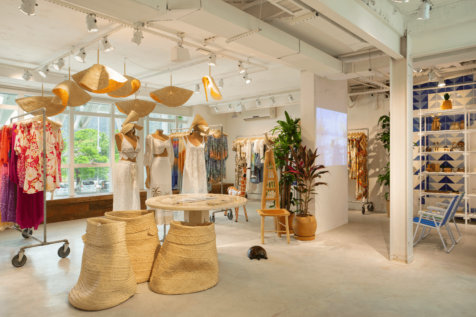 Interior of a clothing boutique. The layout is modern. Three mannequins in the centre of the room are dressed in bohemian, beachy attire. Two beach chairs face a film projection on an otherwise blank wall. There are exposed steel beams and a steel pillar, and ceiling lights with shades that resemble sun hats. Three large, slouchy raffia baskets sit on the cement floor next to a round table. Four racks of clothing in warm colours are dotted around the room. A shelving unit holds jewelry and home decor.