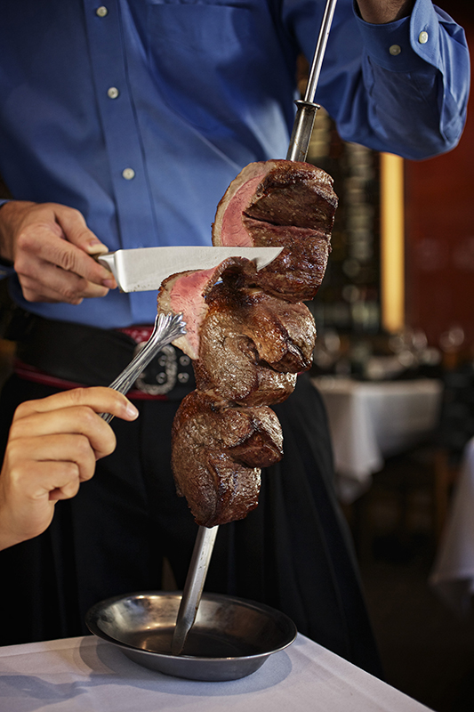 A waiter holds a large skewer of cooked meat at a table. It appears to be beef. The waiter is slicing the meat while a second person uses a tongs to hold the piece of meat that is being cut from the skewer. A second table is visible in the background.