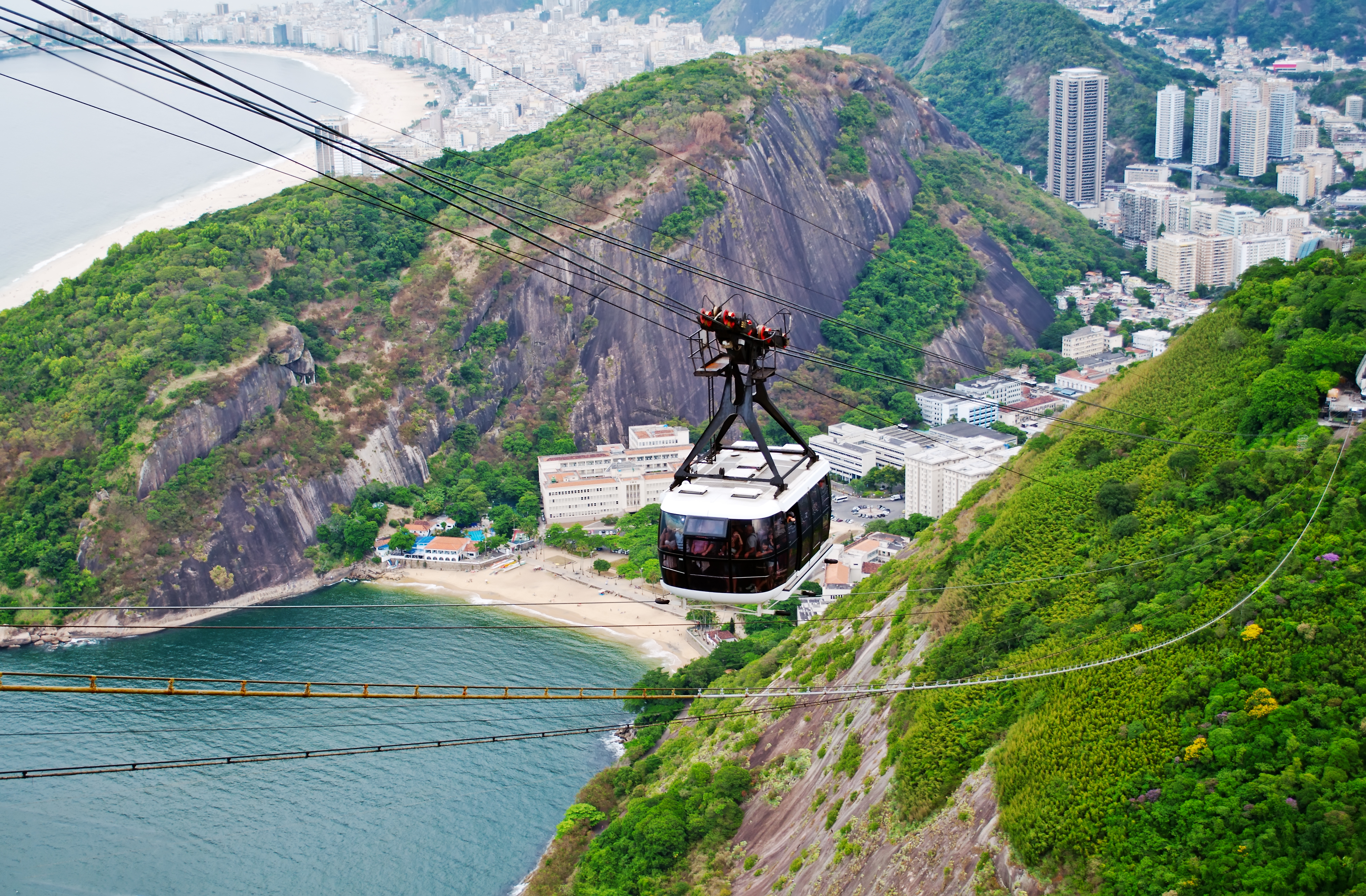 A cable car with floor to ceiling glass walls is suspended, high above the ground, from cables joining a rocky, forested hilltop to something out of frame. Behind the cable car, at sea level, is a beach in a bay between two rocky, forested hills. Numerous multi-story buildings spread inland from the beach.