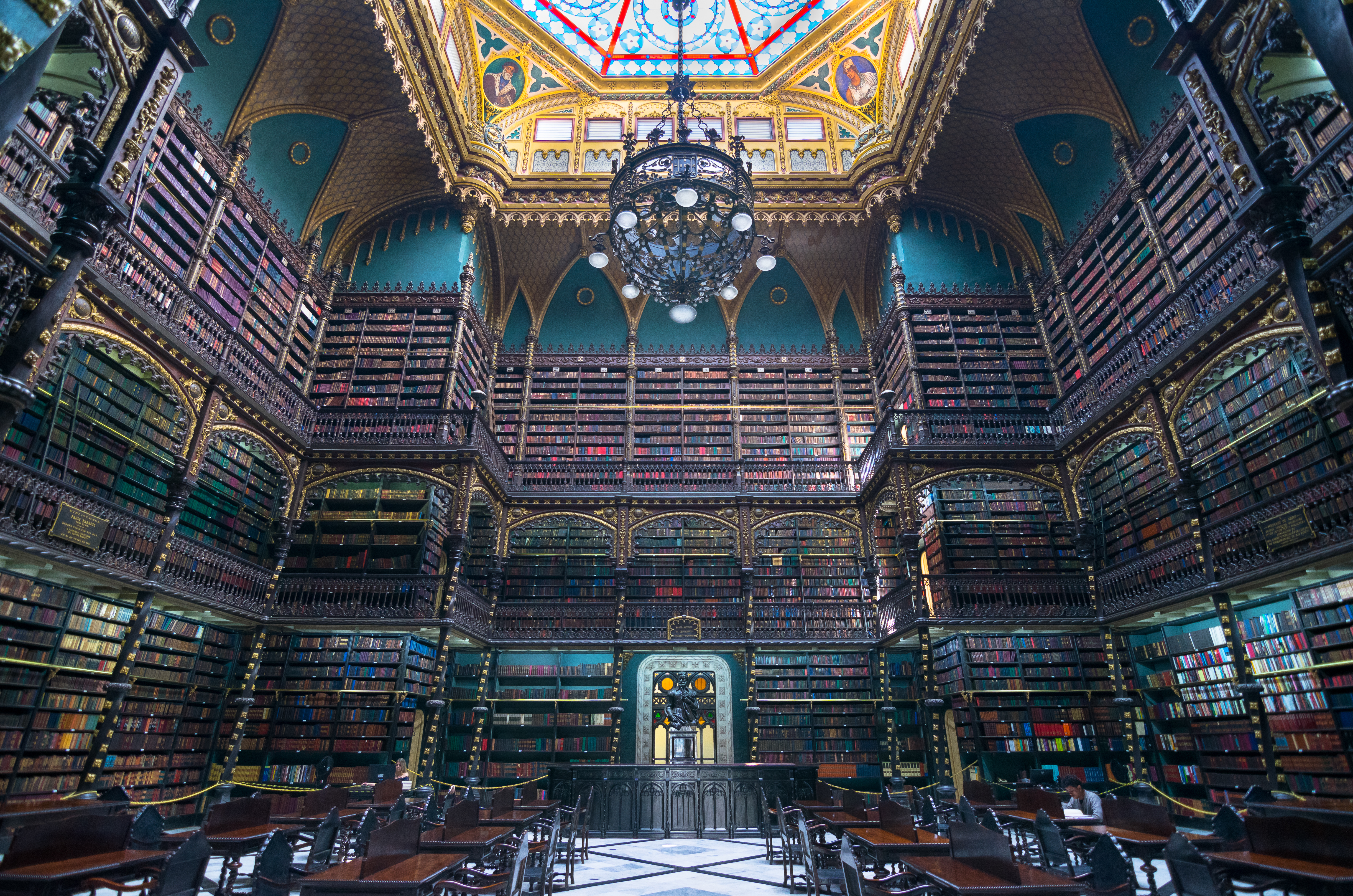 The interior of a library, gilded and decorated in an extravagant style. The walls are densely packed with books over three floors. A chandelier is suspended from the ceiling, which is painted in part with what appears to be gold leaf. The centre of the space is empty other than tables and chairs.