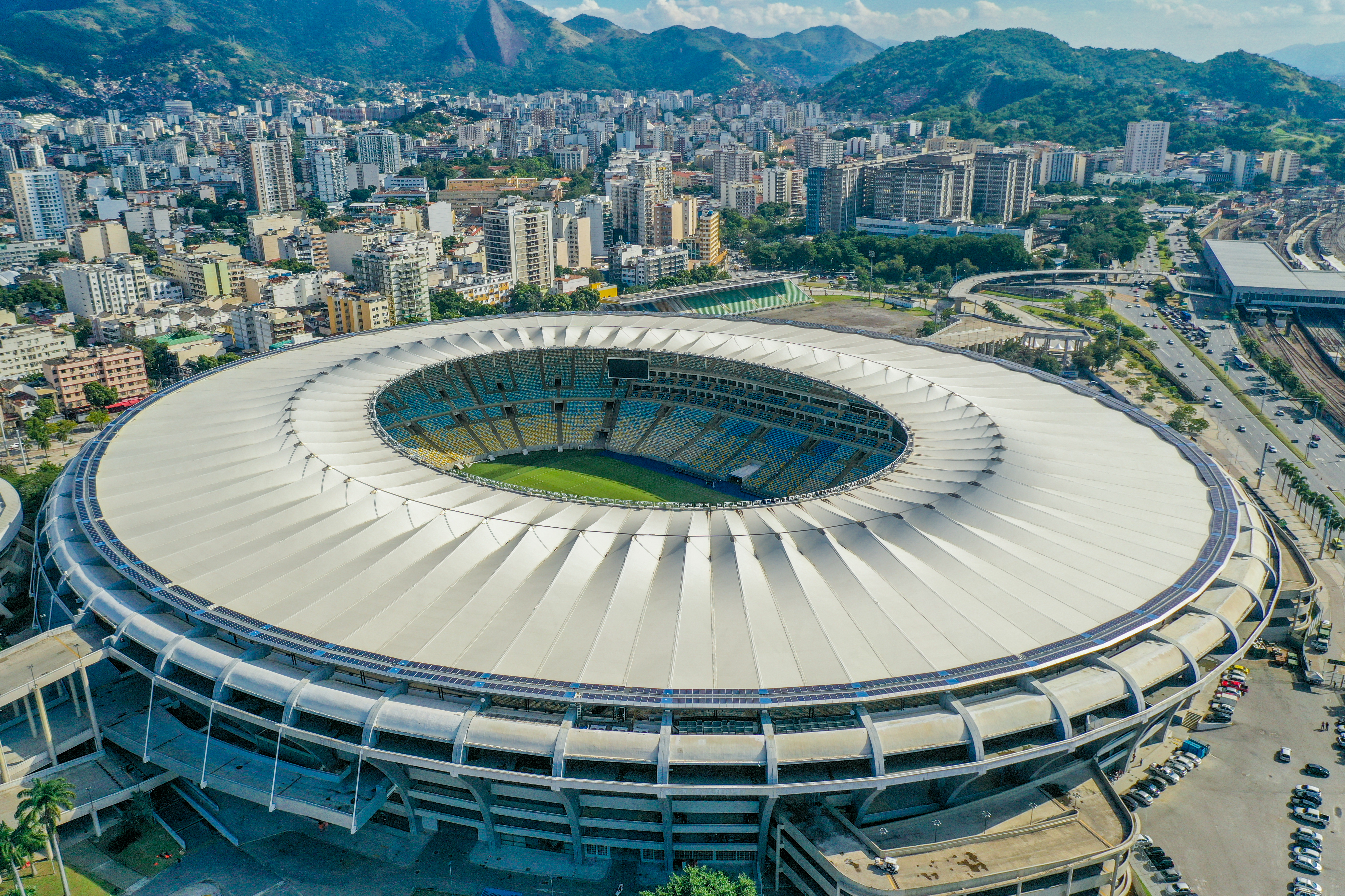 Aerial view of Maracanã stadium. The partially-covered stadium is circular, with a circular gap in the roof directly above the pitch. Behind the stadium sprawls the city of Rio, with many high-rise buildings visible. Rocky, forested mountains are visible in the background.
