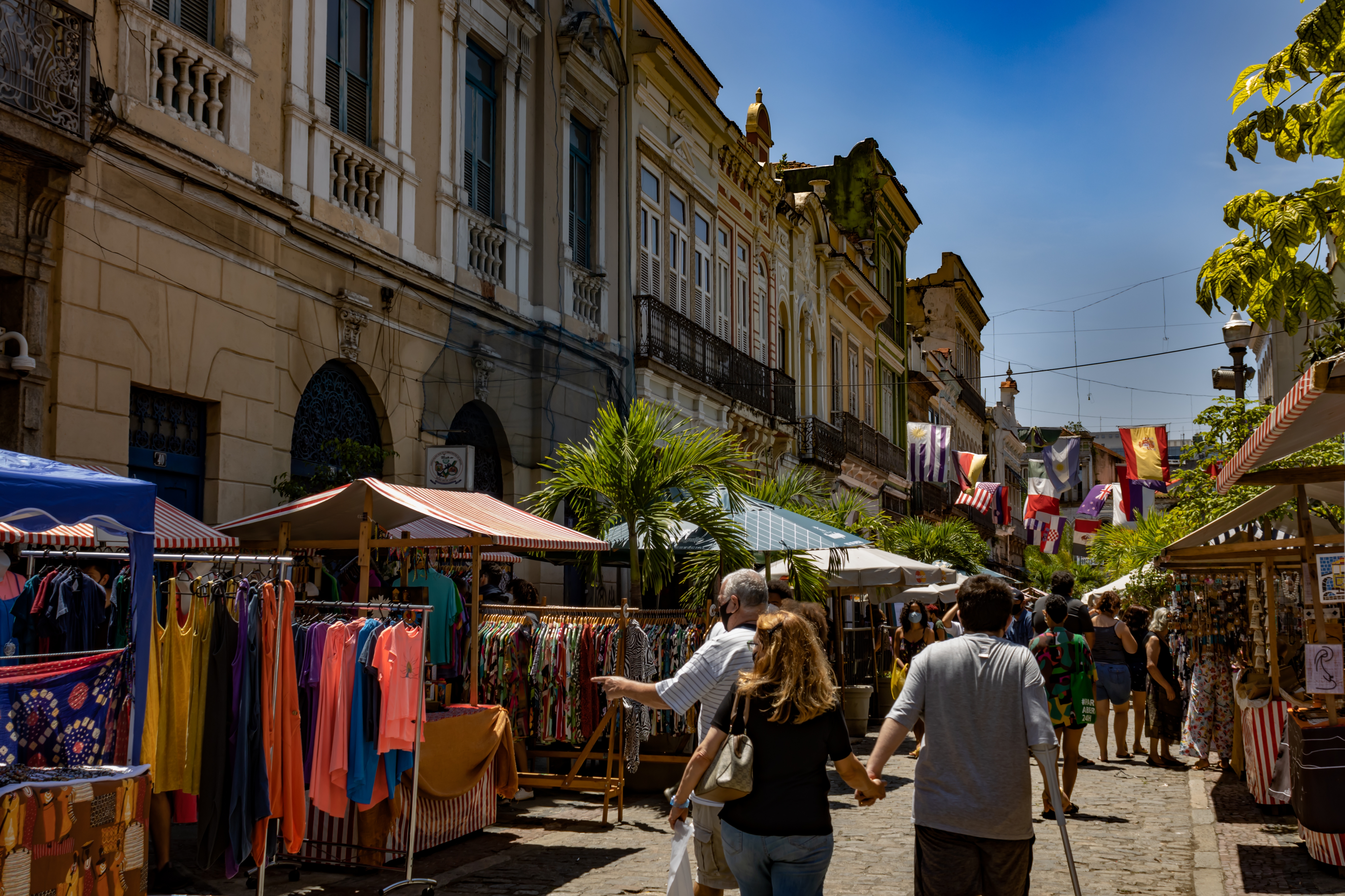 A street market on a tree-lined avenue in sunny Rio. People in summer clothes wander between stalls that are pressed against the bottom of old, terraced stone buildings with cast-iron balconies. The stalls are mostly selling clothing. A number of flags hang above the street, including the Spanish, Mexican and Croatian flags.