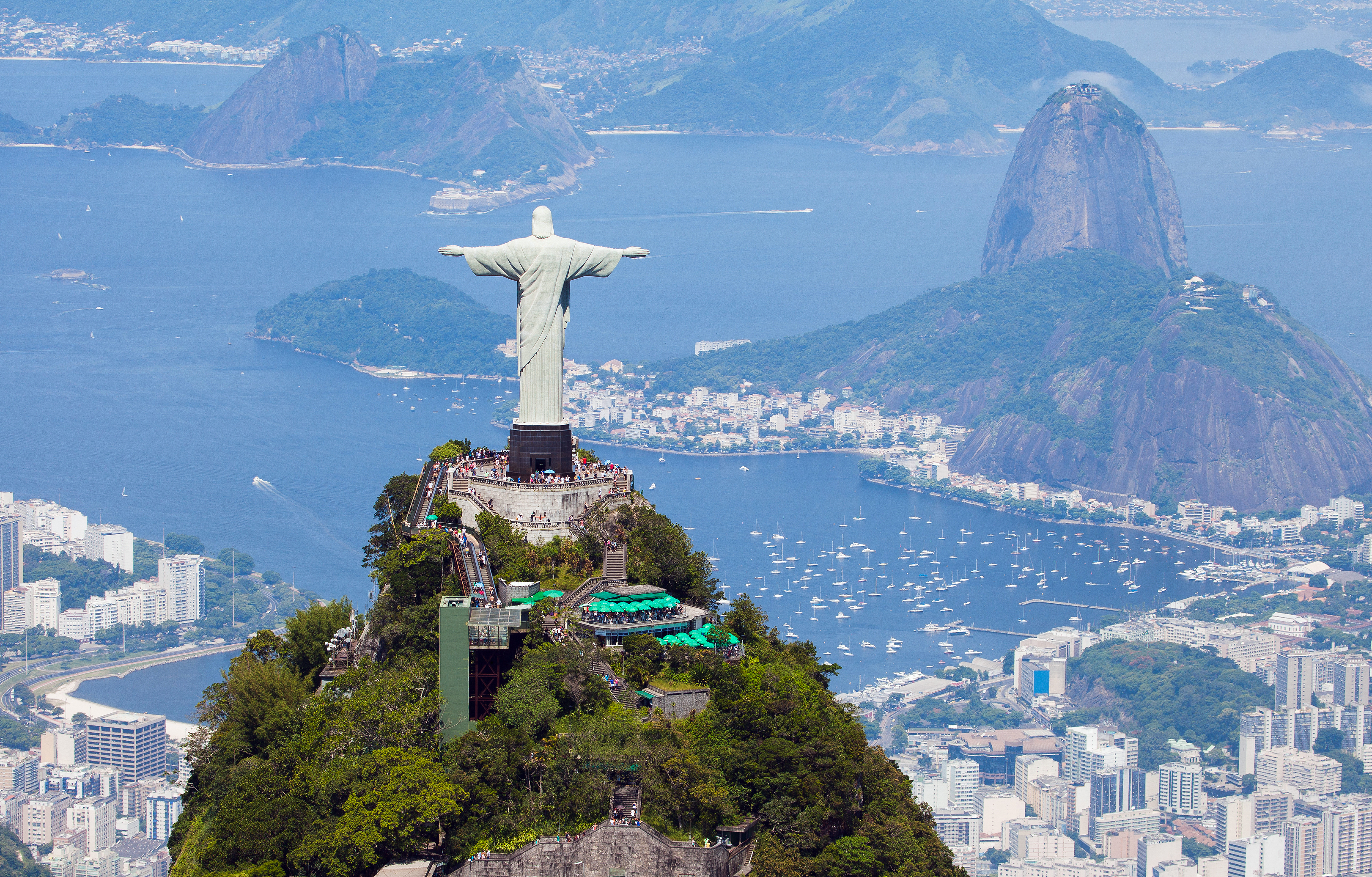 A statue of a male figure with arms outstretched – Rio's Christ the Redeemer statue – stands on top of a rocky outcrop, facing away from the camera. In the background are the rocky Sugarloaf Mountain and a bay lined by skyscrapers.
