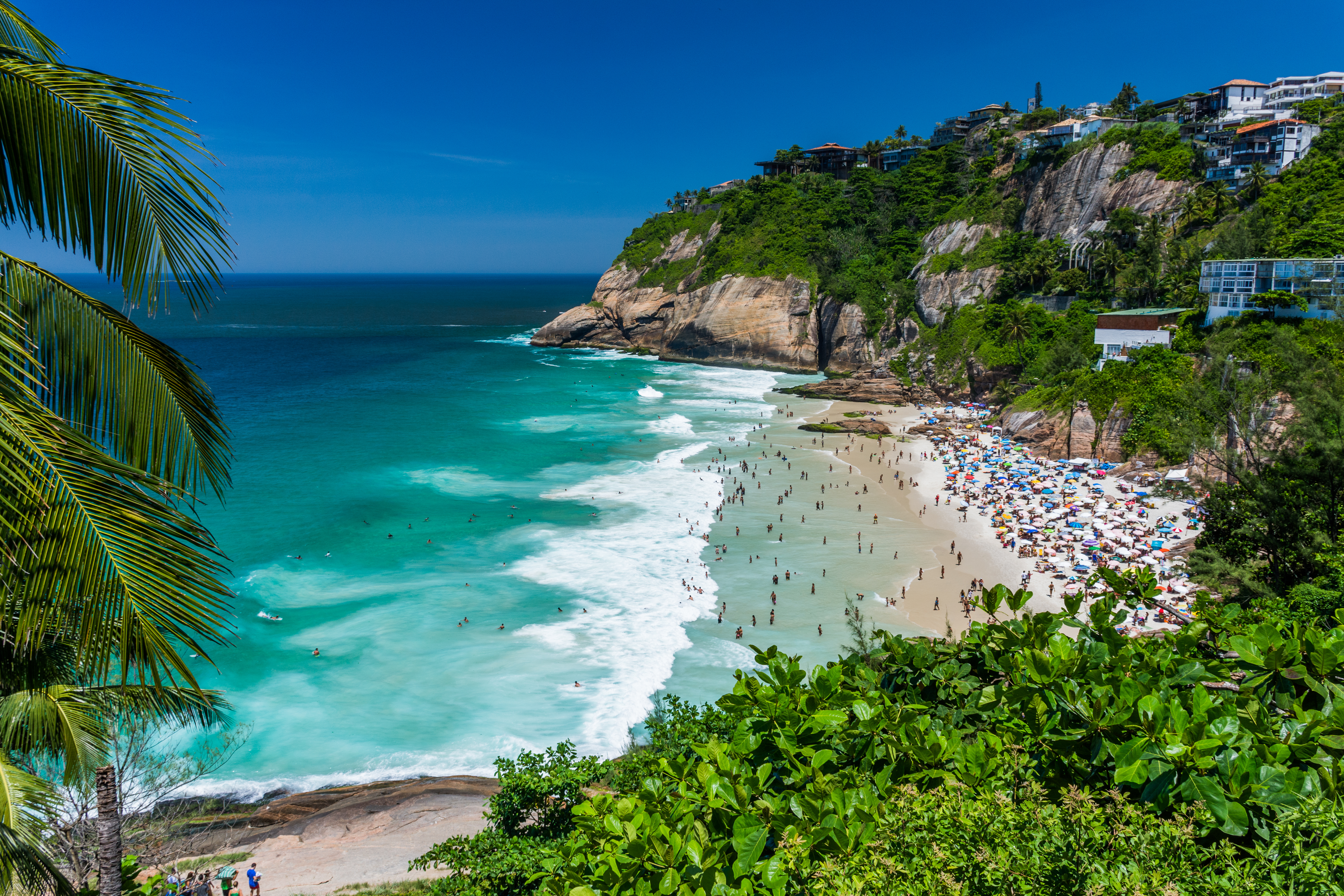 A crowded beach is visible through a gap in the trees. The beach is surrounded by rocky, tree-topped cliffs. Residential buildings dot the cliffs. People are scattered through the water, and appear to be swimming. Gentle foamy waves are visible where the water meets the beach.