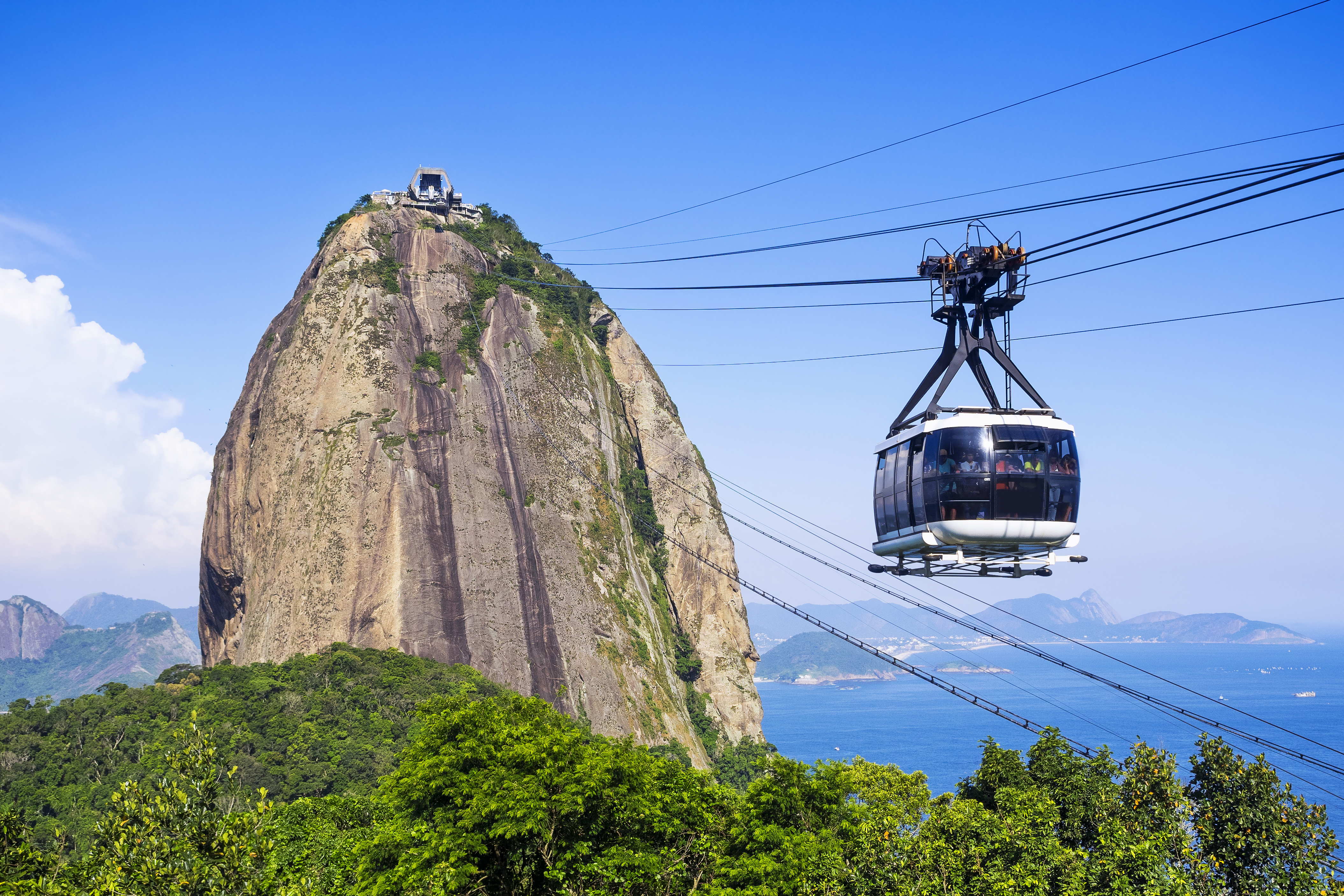 A glass-sided cable car is suspended from cables above dense leafy forest. The cable car appears to be moving towards the summit of Rio’s Sugarloaf Mountain, a tall but narrow mountain jutting out of the forest. The sea is visible in the background.