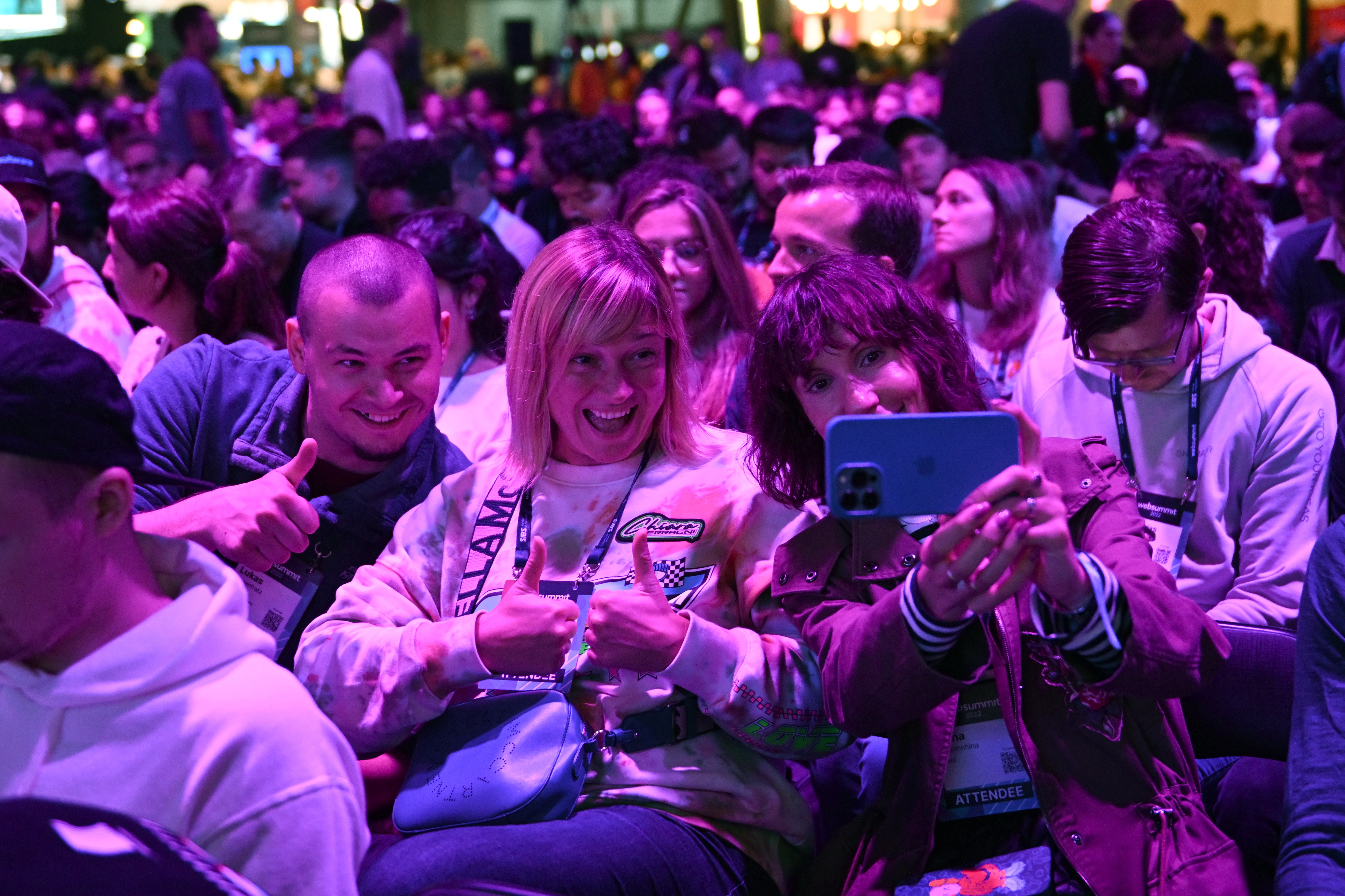 Three attendees at the front of a large crowd of seated people lean towards each other. One of these people is holding a mobile phone. The other two people are giving thumbs up. The three appear to be taking a selfie.