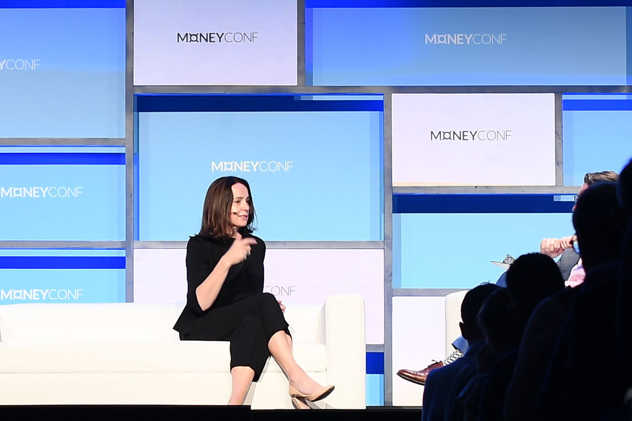 A person (Square CFO Sarah Friar) sits on a couch. The person's legs are crossed and they're gesturing with their right hand. The person is wearing a headset mic, and appears to be speaking. On the wall behind the person, the MoneyConf logo appears in several places.