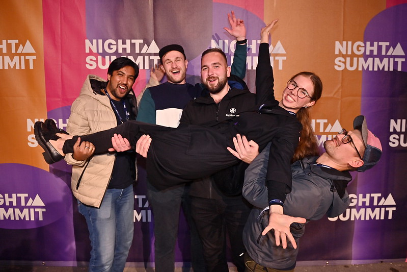 Five people pose for a photo in front of a wall on which the Night Summit logo appears several times. The people are smiling. Four of the people are carrying the fifth person, who is stretched out across them.
