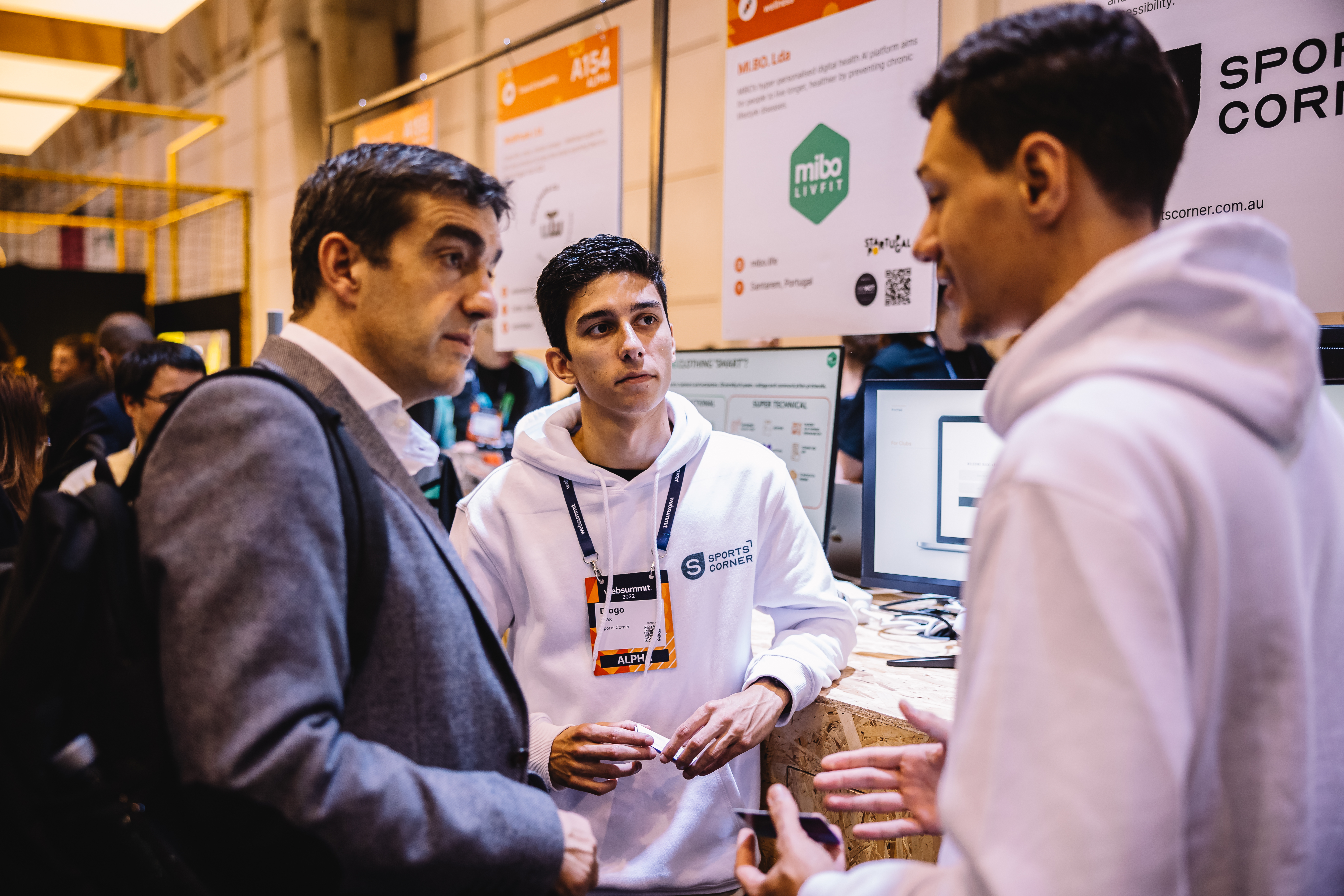 Three people standing. Two of the people face the third person, who appears to be speaking. This speaking person is gesturing with their hands. The speaker and one of the other people are wearing hoodies. The third person is wearing a suit jacket and carrying a backpack over one shoulder. Behind the people are a computer screen and several exhibition boards detailing startups' information.