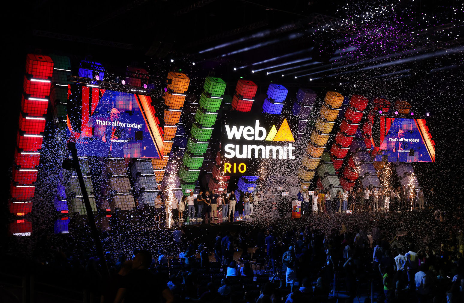 A large stage viewed over the silhouetted heads of a packed audience. A line of people stands on the stage, facing towards the audience. Confetti fills the air. A large Web Summit Rio logo hangs in the center of the stage. Two screens on either side of the logo read 'That's all for today!'