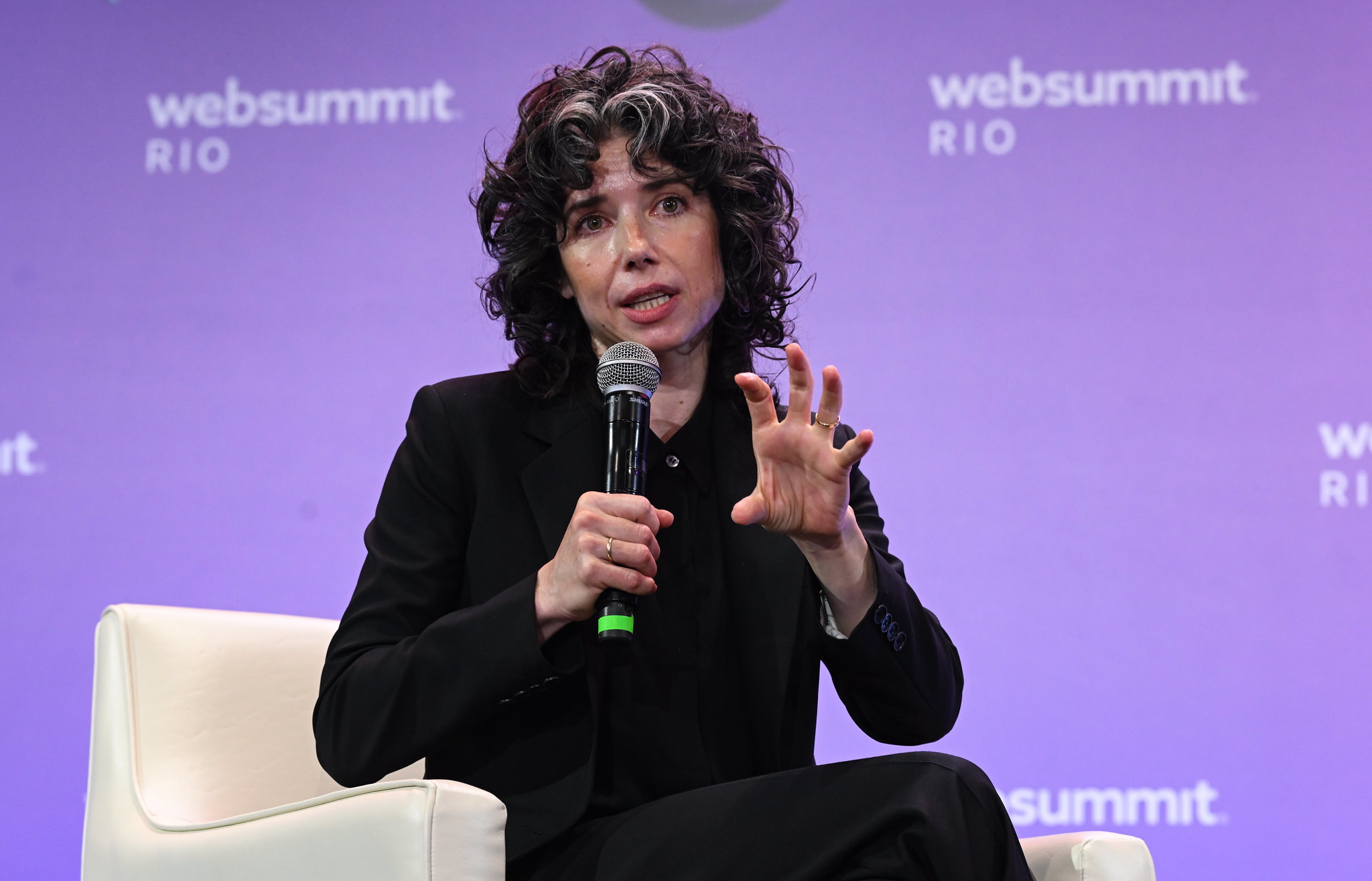 A person (Signal president Meredith Whittaker) sits in an armchair. They hold a microphone in their right hand and are gesturing with their left. They appear to be speaking. On the wall behind them, the Web Summit Rio logo is visible in several places.