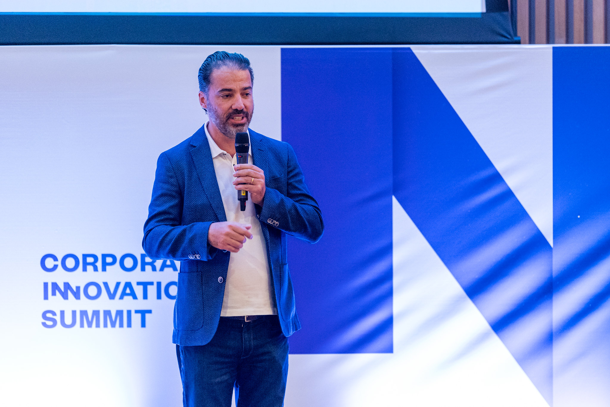 A person stands in front of a wall that reads 'Corporate Innovation Summit'. They're holding a microphone in their left hand and are gesturing with their right. They appear to be speaking.