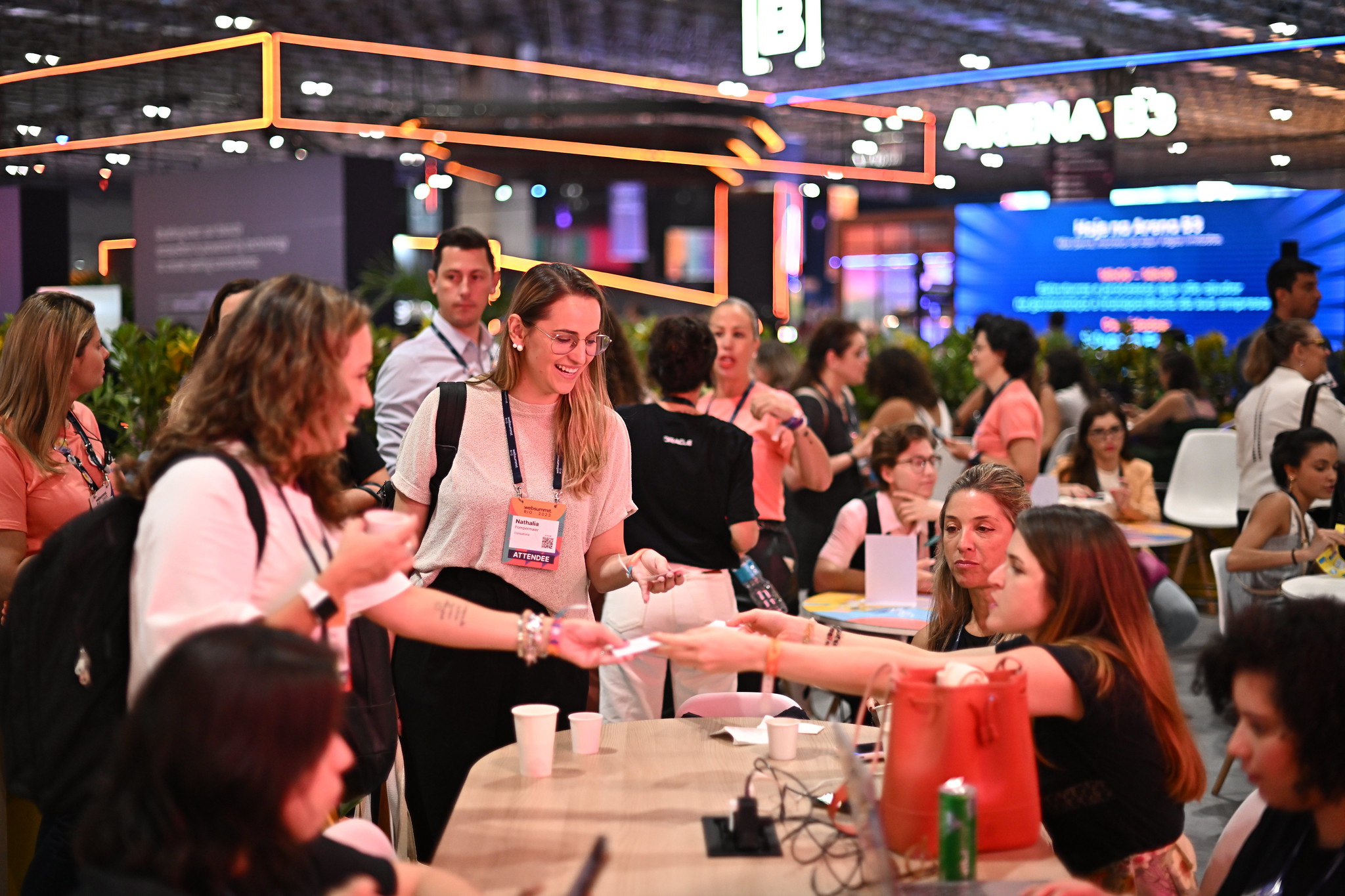 A busy lounge area. People are seated at round wooden tables, and standing in groups. People are smiling and chatting. Visible in the background, behind a leafy hedge, are large stands on an exhibition floor. This is the Women in Tech Lounge at Web Summit Rio.