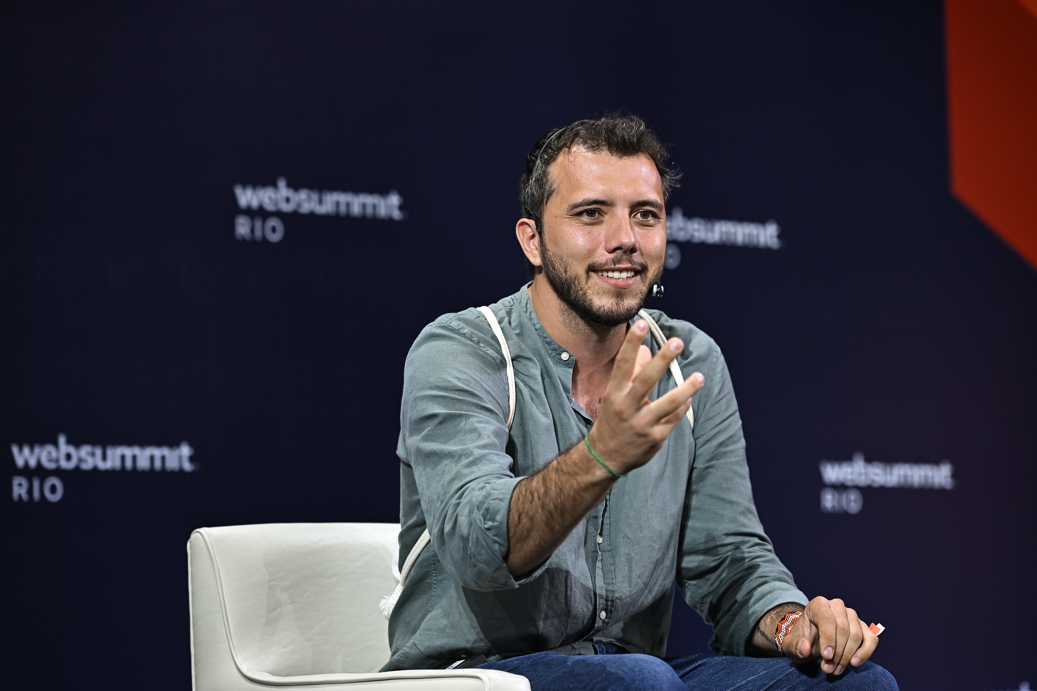 A person (Movimento Bem Viver co-founder Thiago Ávila) sits in an armchair. Their left arm rests on their knee. They gesture with their right hand, reaching towards the audience, which is out of shot. They are wearing a headset mic.