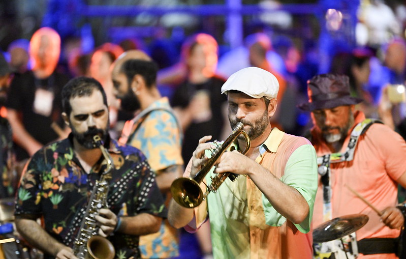 A person playing saxophone, a person playing trumpet and a person playing drums. All three are standing. Behind the three musicians, a crowd of people mills about. This is Night Summit at Web Summit Rio 2023.