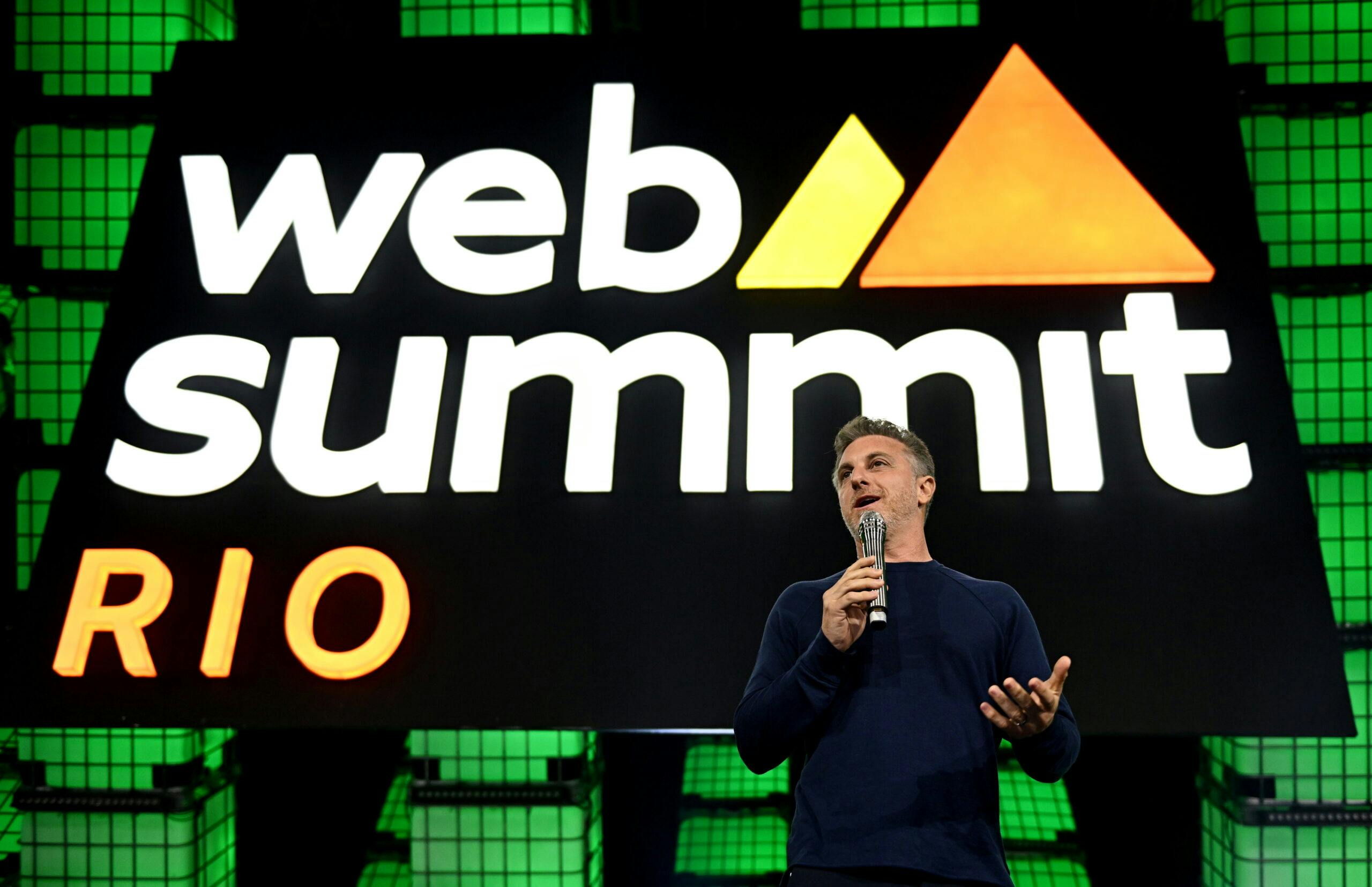 A person (TV star Luciano Huck) in front of a large sign that reads 'Web Summit Rio' and features the Web Summit Rio logo. The person is holding a microphone in their right hand and is gesturing with their left hand. They appear to be speaking.
