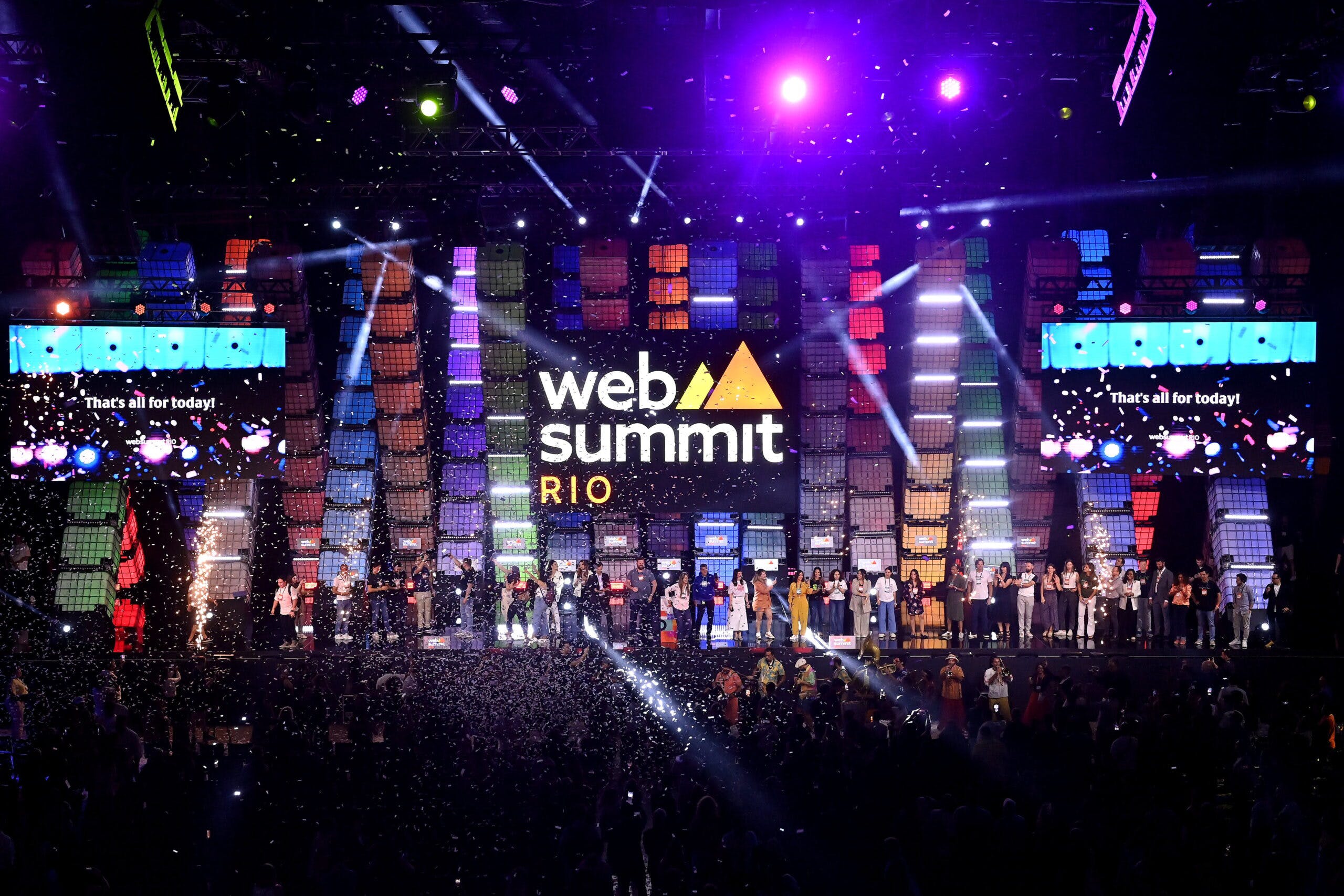 A large stage viewed from a distance over the silhouetted heads of the audience. A long line of people stands on stage, facing the audience. In front of the stage, a group of musicians – including drums, trombones, trumpets, and a sousaphone – appear to be playing. The air is filled with confetti. A large Web Summit Rio logo hangs over the heads of the people on stage. Two large screens on either side of the logo bear the message 'That's all for today!'.