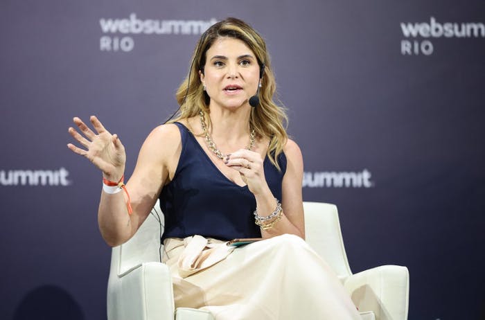 A person (Falconi CEO Viviane Martins) sits in an armchair. Viviane is wearing a headset mic and gesturing with both hands. Viviane appears to be speaking. The Web Summit Rio logo is visible in several places on the wall behind Viviane.