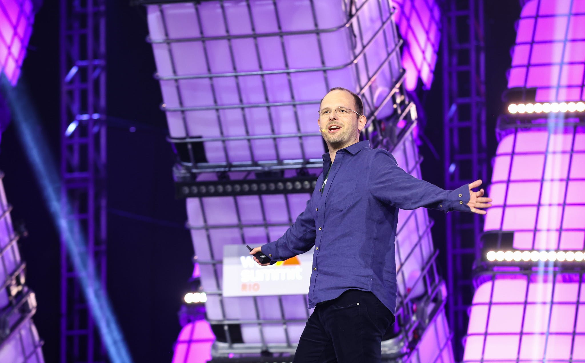 A person (GitHub CEO Thomas Dohmke) walks across a stage. Pictured from the knees up, Thomas is wearing a headset mic and holding a presentation clicker in one hand. Thomas's arms are outstretched. Thomas appears to be speaking.