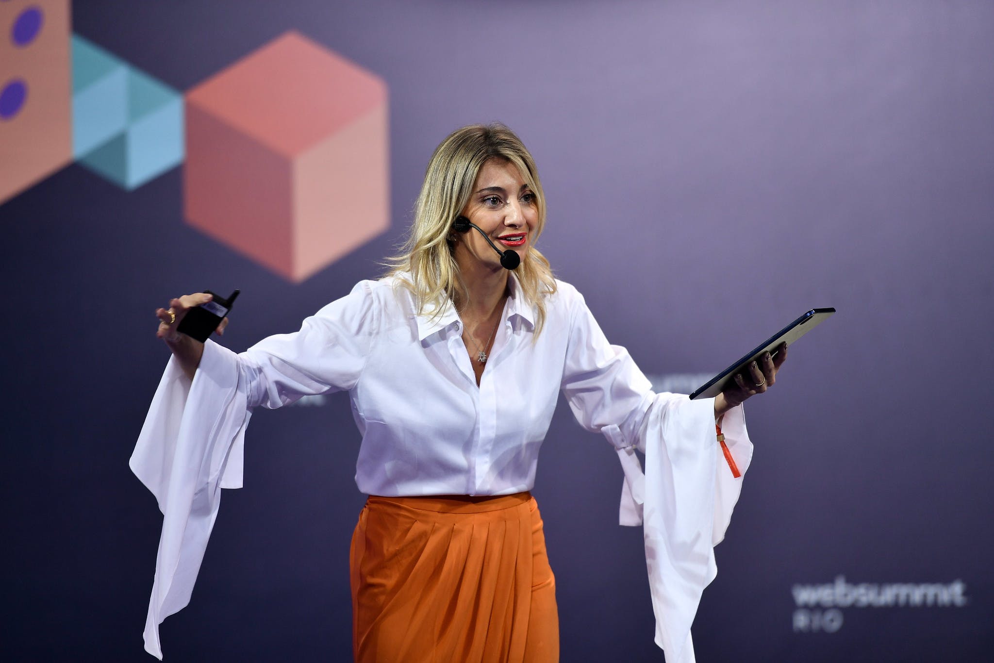 Monique Lima, co-founder and CEO, Mimo Live Sales, stands on stage with arms outstretched while speaking. Monique wears a white blouse and orange skirt.